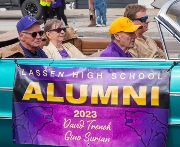 Lassen High School alumni parade with four older individuals sitting in a convertible car, with a purple and gold banner displaying the school name, year 2023, and names David French and Gino Surian.