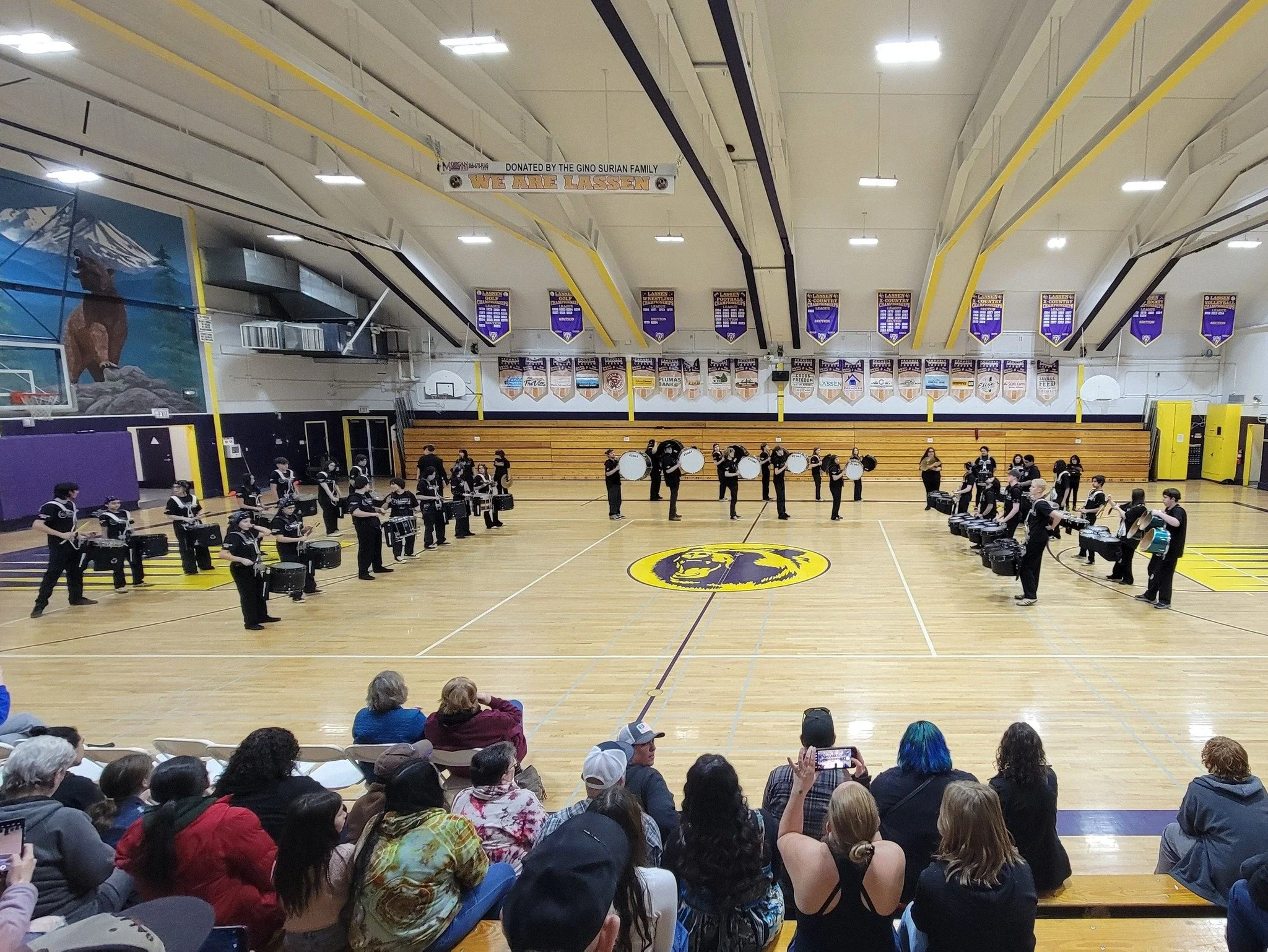 A band performance in a gymnasium with members playing drums and percussion, audience seated on bleachers, banners hanging on walls, and a logo in the center of the court.