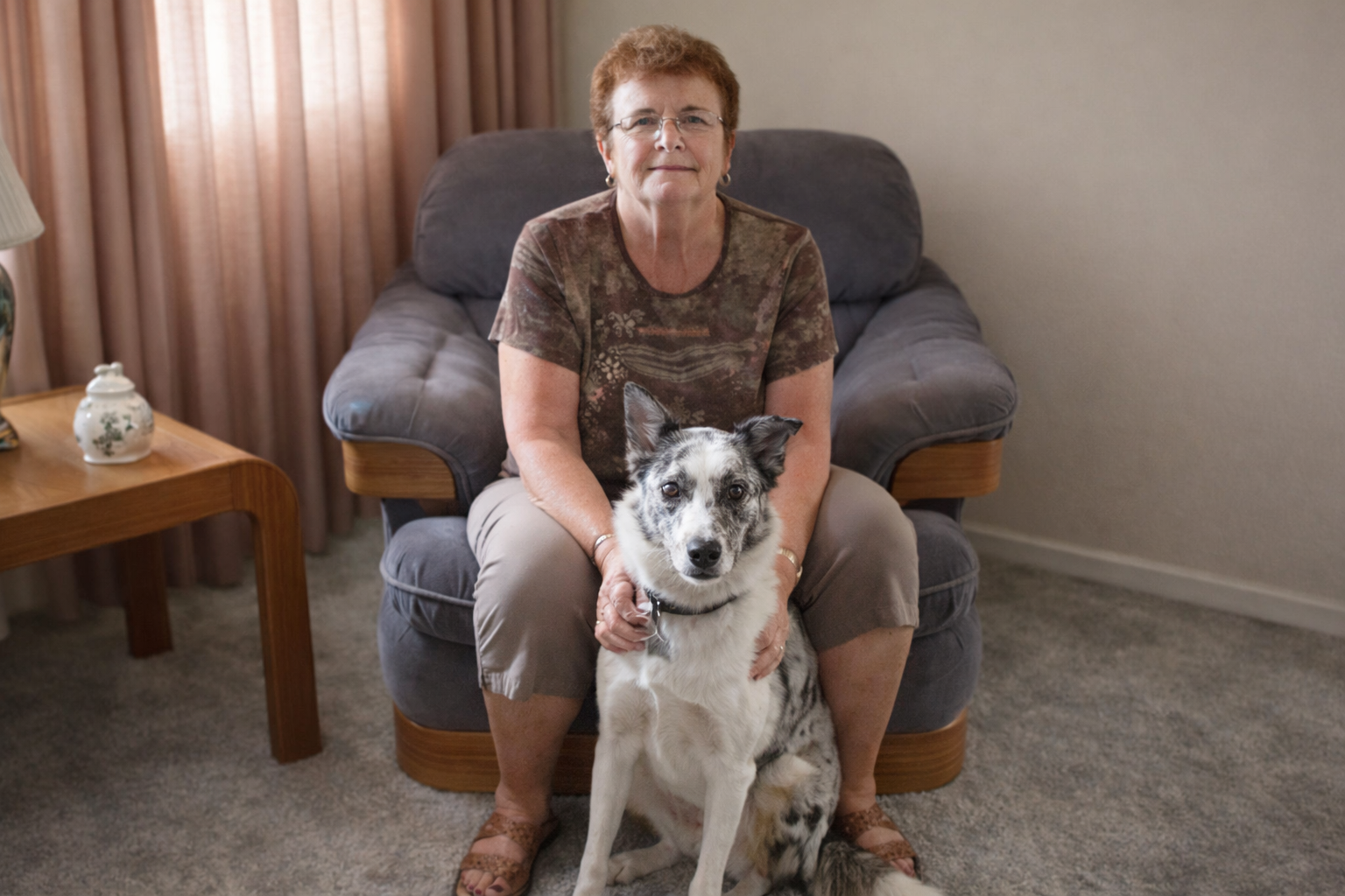 An older woman with short red hair and glasses sitting on a plush gray armchair in a living room, holding a black and white Australian shepherd dog with a black collar, in front of her.