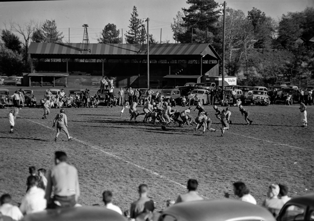 Vintage black and white photo of a football game on a grassy field with spectators sitting and standing along the sidelines, and cars parked behind a small bleacher in the background.