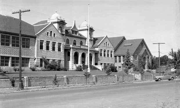 Historical black-and-white photo of a large institutional building with domed towers, multiple windows, and a central staircase, located along a street with power poles and a parked car.