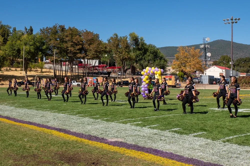 Cheerleaders performing on a football field during a game, with balloons and trees in the background on a sunny day.