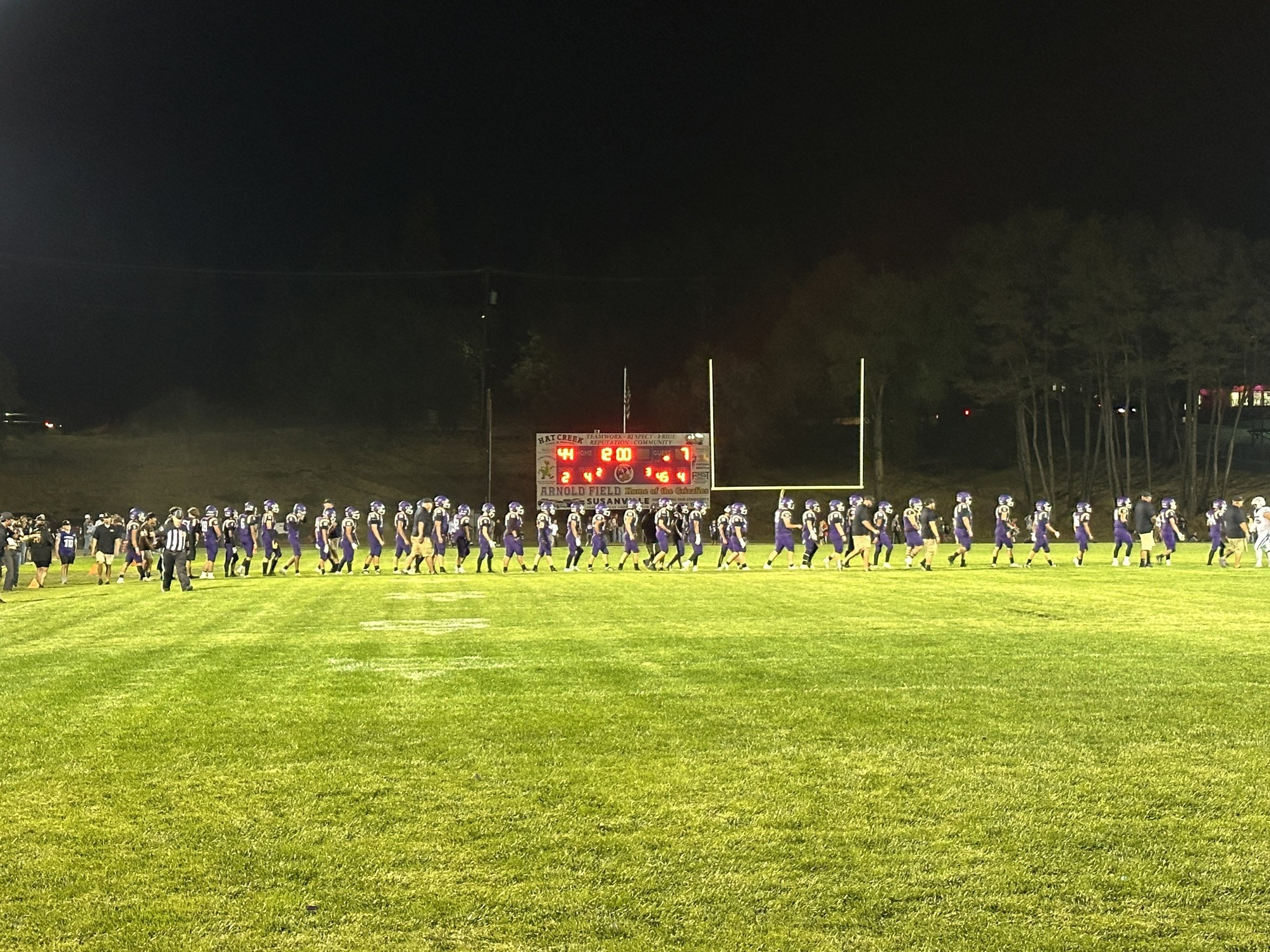 High school football team walking on the field at night after a game under bright stadium lights, with a scoreboard showing a final score of 44-7.
