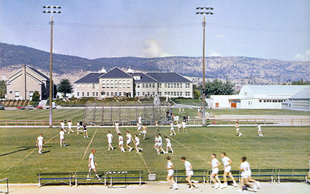 A group of people in white sports uniforms warming up or practicing on a grassy field, with bleachers, buildings, and hills in the background.