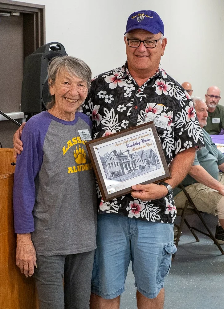An elderly woman and a middle-aged man smiling and posing together, with the man holding a framed drawing of a historic building. The woman wears a Lafayette Alumni T-shirt, and the man wears a floral Hawaiian shirt and a blue cap. Several people are seated in the background at an indoor event.
