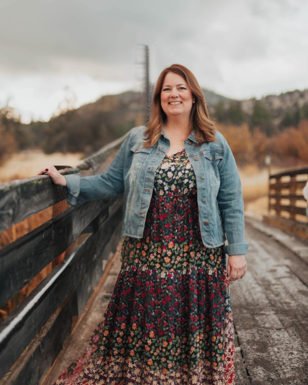 A woman with shoulder-length brown hair, smiling, wearing a denim jacket over a floral dress, standing on a wooden bridge outdoors with a mountainous landscape in the background under a cloudy sky.