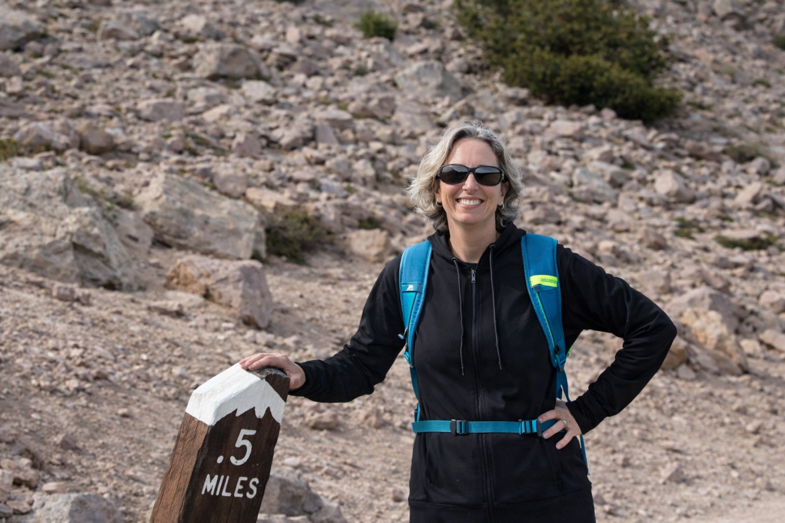 A smiling woman with gray hair wearing sunglasses, a black jacket, and a blue backpack, standing outdoors on a rocky trail next to a sign that reads '.5 MILES.'