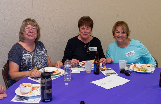 Three women sitting at a table with blue tablecloth, having food and drinks, smiling for the camera, in a casual setting.