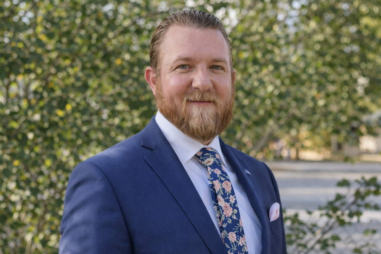 A man with a full beard and mustache wearing a blue suit, white shirt, floral tie, and a pocket square, smiling outdoors with green trees in the background.