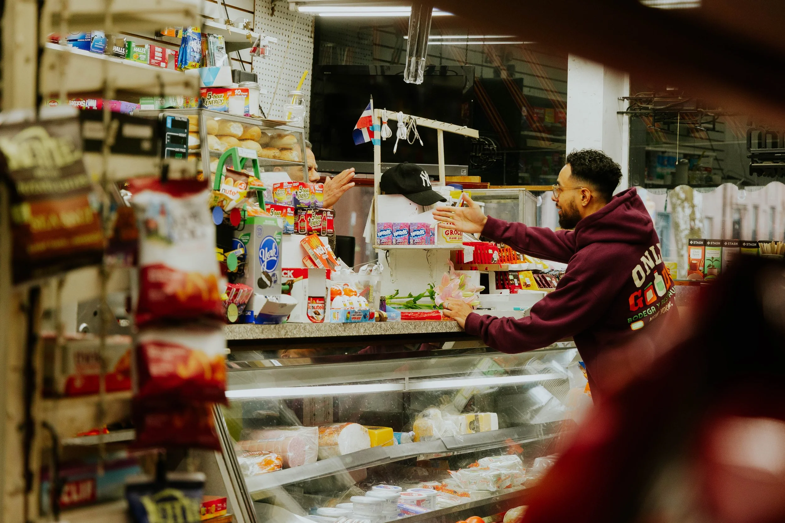A young man wearing glasses and a maroon hoodie interacts with a cashier at a grocery store counter, which displays various snacks, candies, and dairy products. The cashier is behind the counter, reaching out to hand over a product.