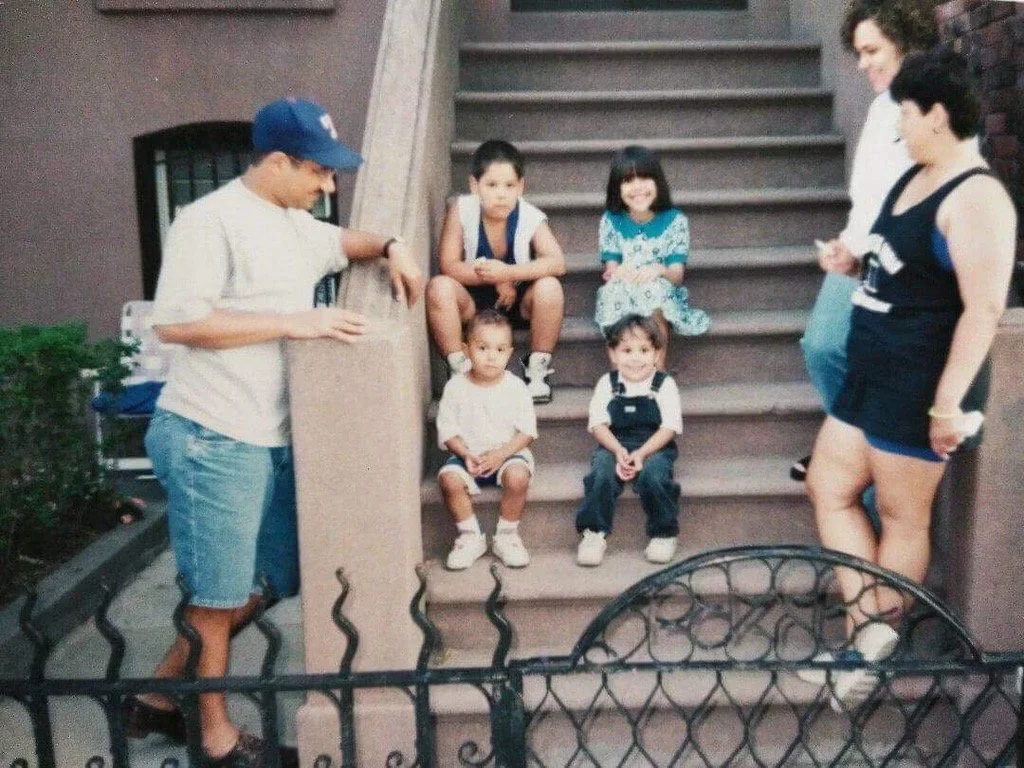 A group of seven children and two adults sitting on and near a stairway outdoors. Four children are seated on the stairs, with two older kids standing to the right and left of the stairs. The children appear to be of different ages and are dressed casually. The adults, a man wearing a baseball cap and a woman in a black tank top and shorts, are standing nearby.
