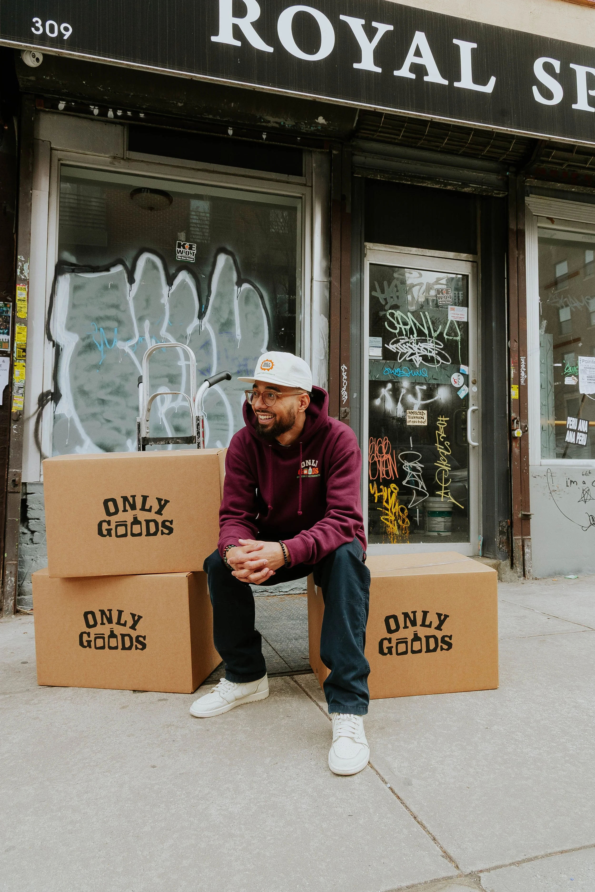 A man sitting on a cardboard box with 'ONLY GOODS' printed on it, outside a graffiti-covered storefront with a glass door and windows, wearing a white cap, glasses, a maroon hoodie, dark jeans, and white sneakers.