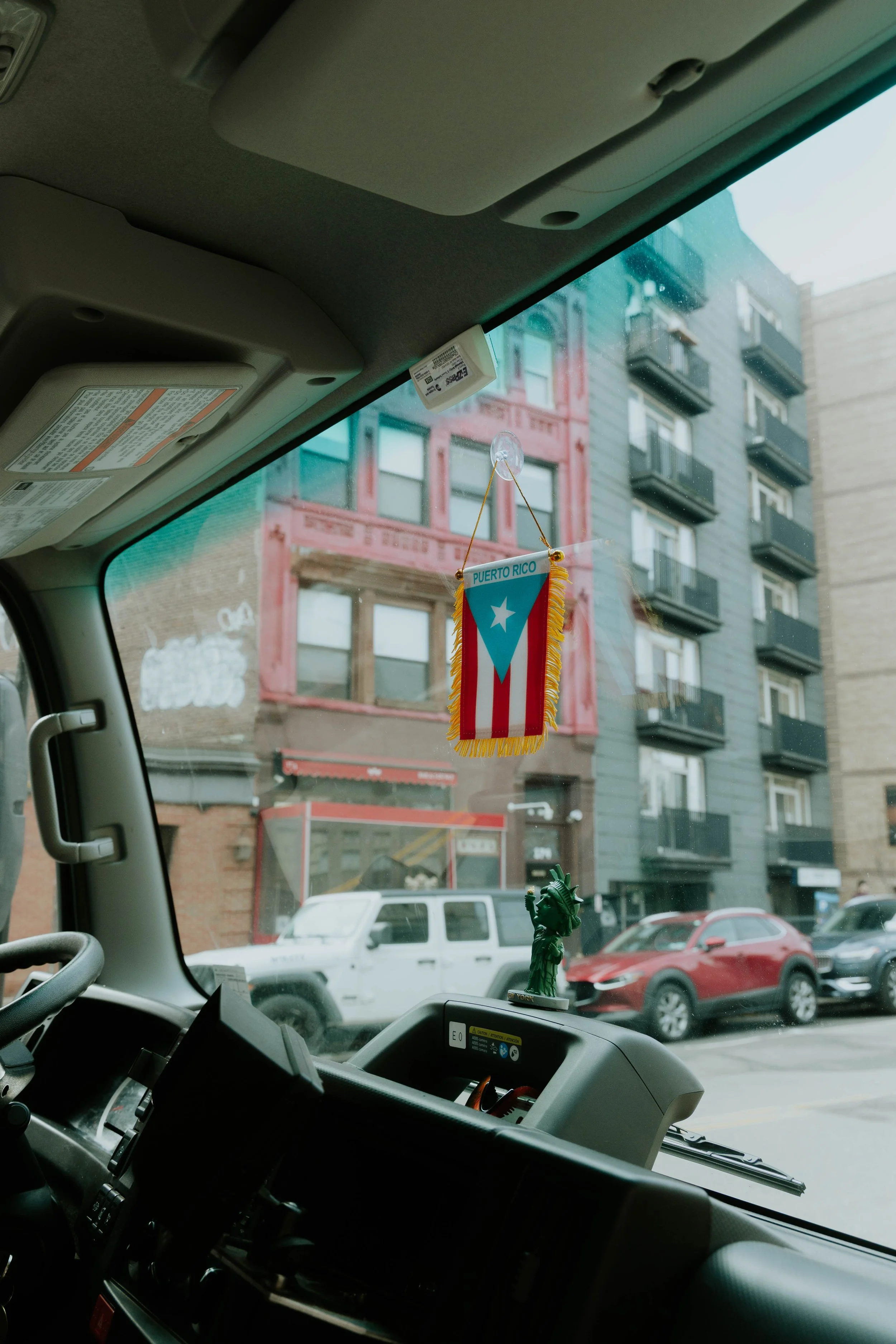 Inside a vehicle, with a Puerto Rican flag decoration hanging from the rearview mirror and a small Statue of Liberty on the dashboard. Outside the window, there are buildings and parked cars.