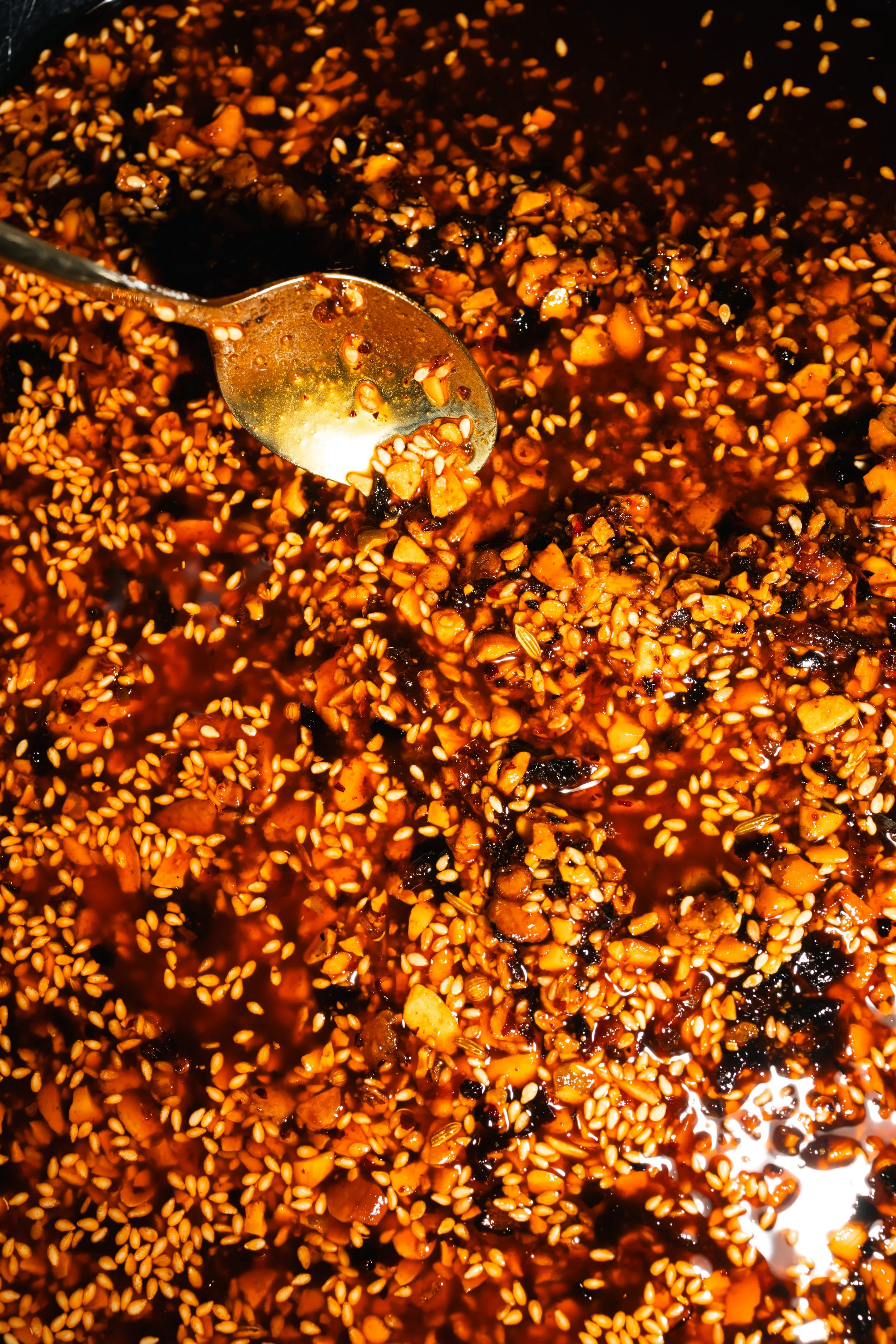 Close-up of a spoon in a mixture of chopped ingredients and sesame seeds with a reddish-brown liquid, likely a sauce.
