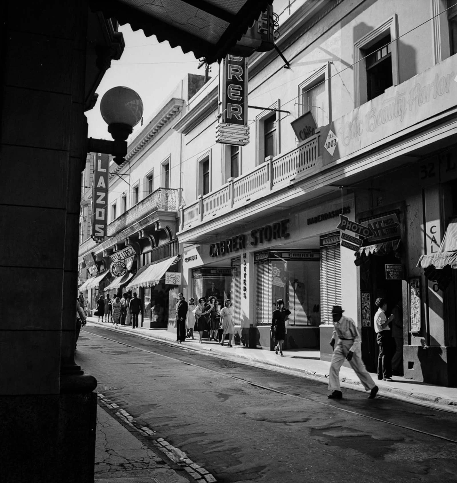 Black and white photo of a vibrant street scene in a city, with people walking along the sidewalk in front of shops and stores, including a laser and barber store, with signs and awnings, and an older man walking across the street.