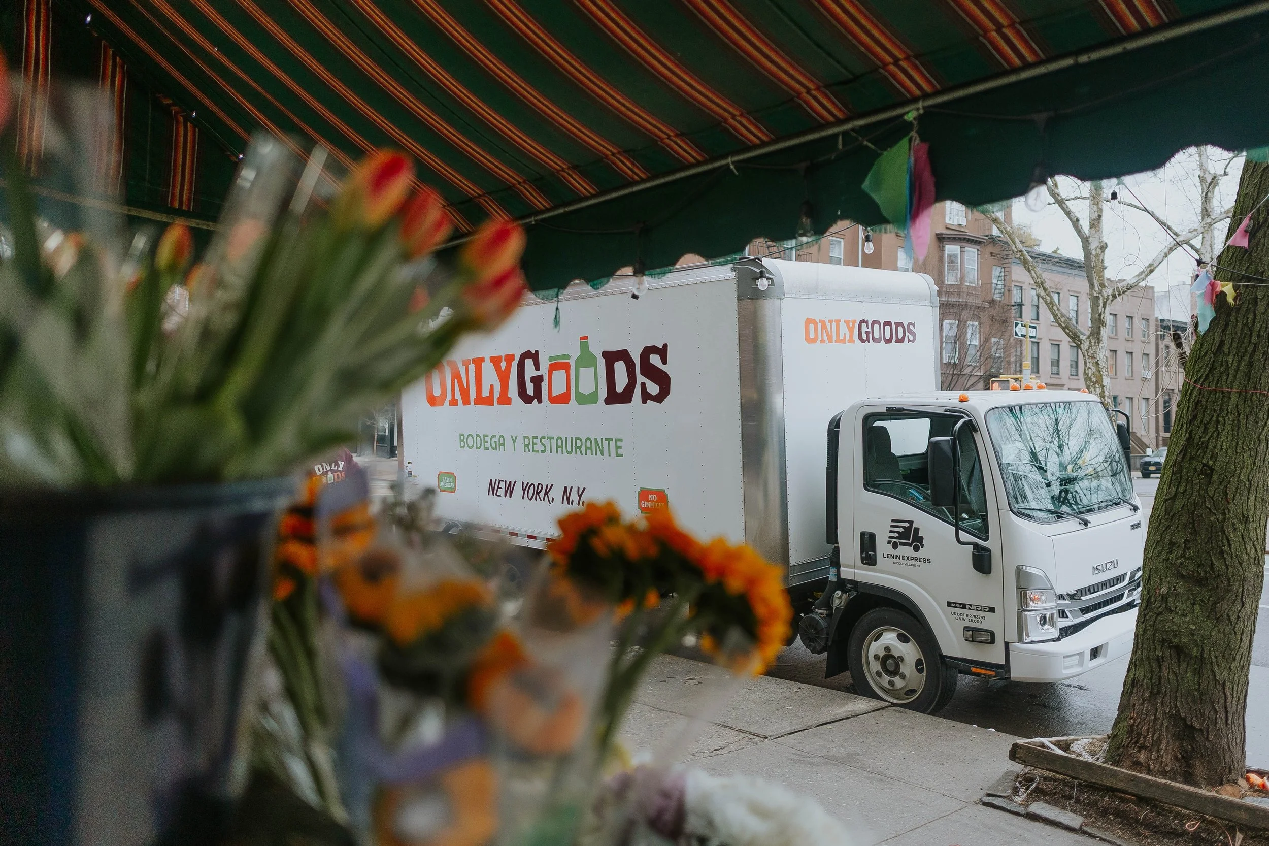 A white delivery truck with 'ONLYGOODS' logo parked on a city street, viewed from behind some blurred flowers in the foreground, with a tree and brick buildings in the background.