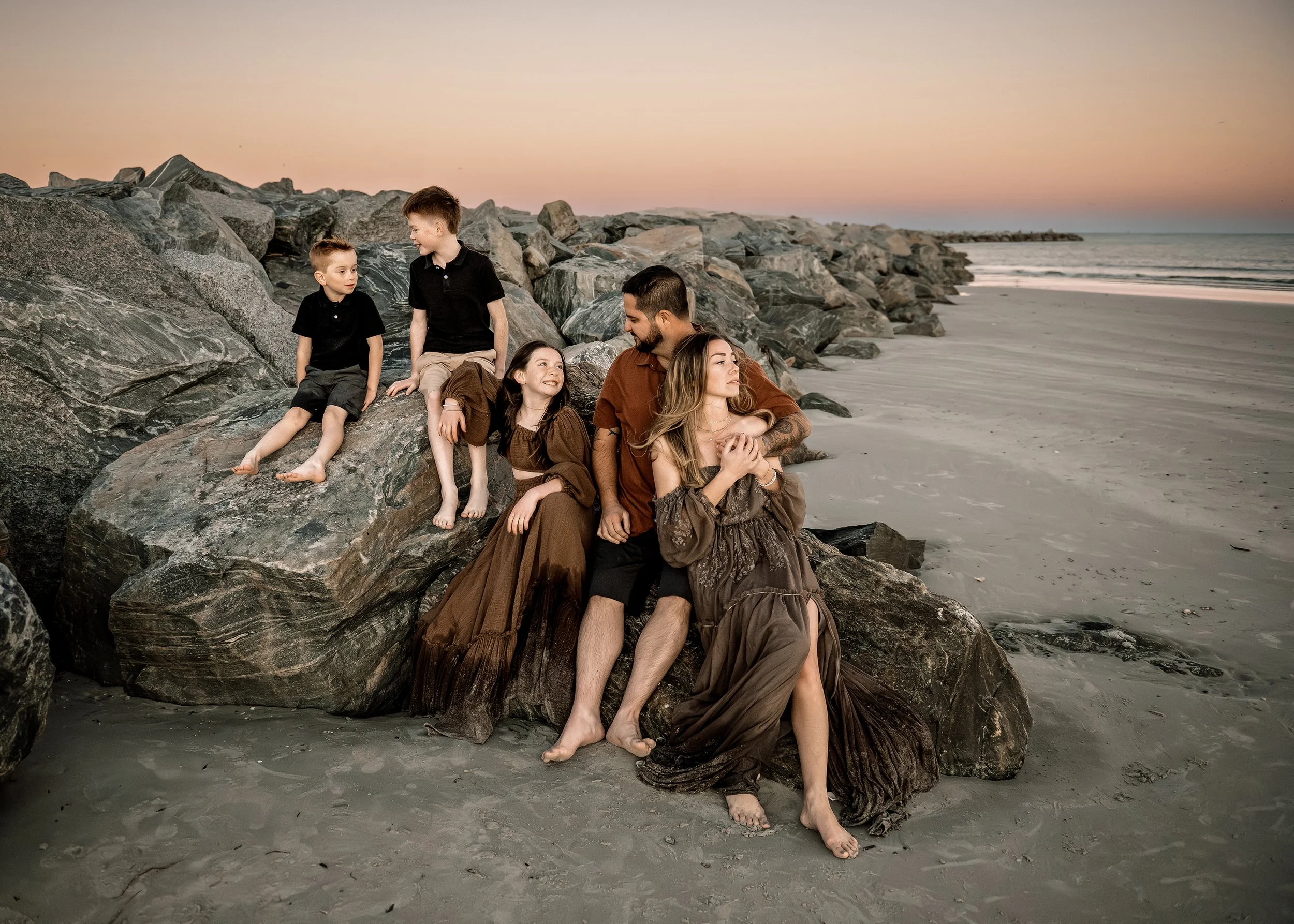 Family sitting on rocks at the beach during sunset, with two women and three children.