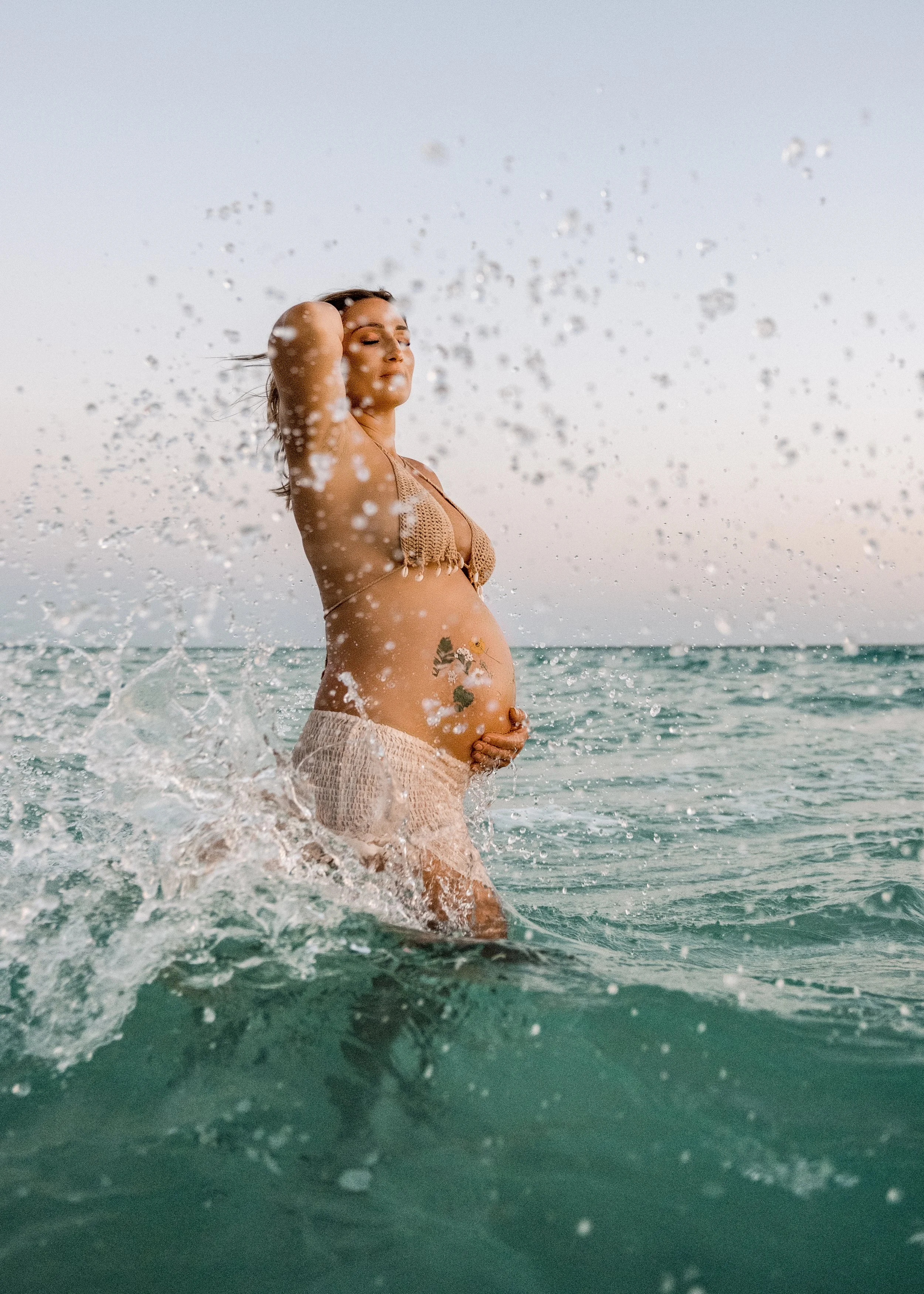 A pregnant woman standing in the ocean, wearing a beige bikini, with her eyes closed and one hand on her stomach. Water splashes around her.