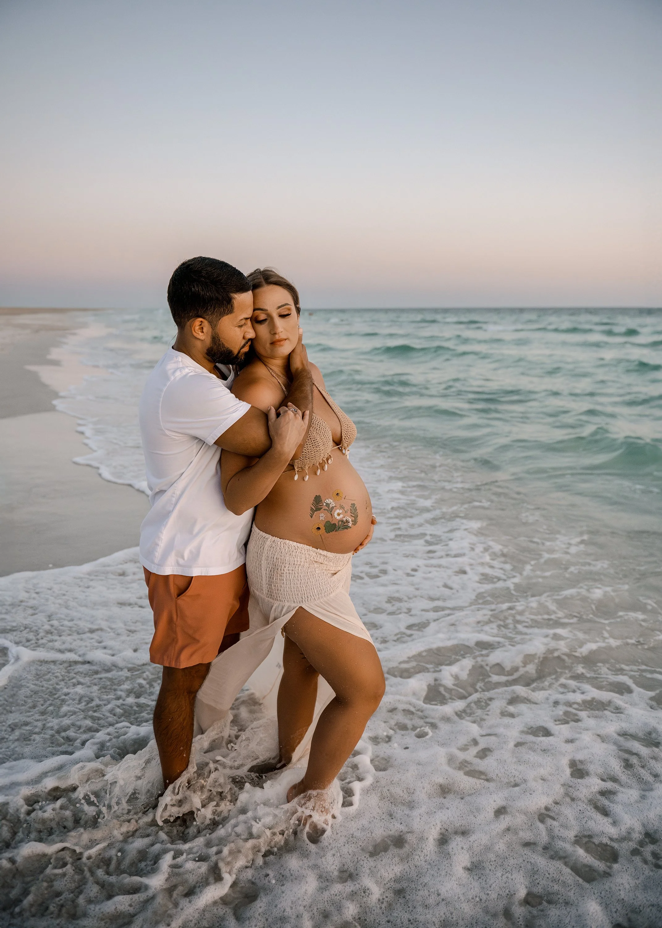 A couple stands in the shallow surf at the beach, embracing and touching foreheads. The woman is pregnant, with a tattoo on her belly, wearing a beige skirt and top. The man wears a white shirt and orange shorts. The sky is pastel, indicating sunset.