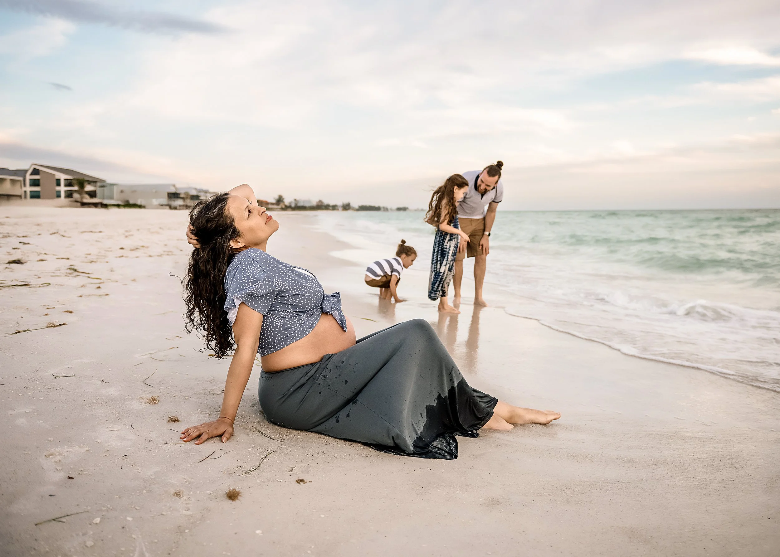 A pregnant woman sitting on the beach with water on her legs, while her family, including a man, a young girl, and a young boy, are near the shoreline. The woman has long dark curly hair, and the background shows a beach house and a cloudy sky.