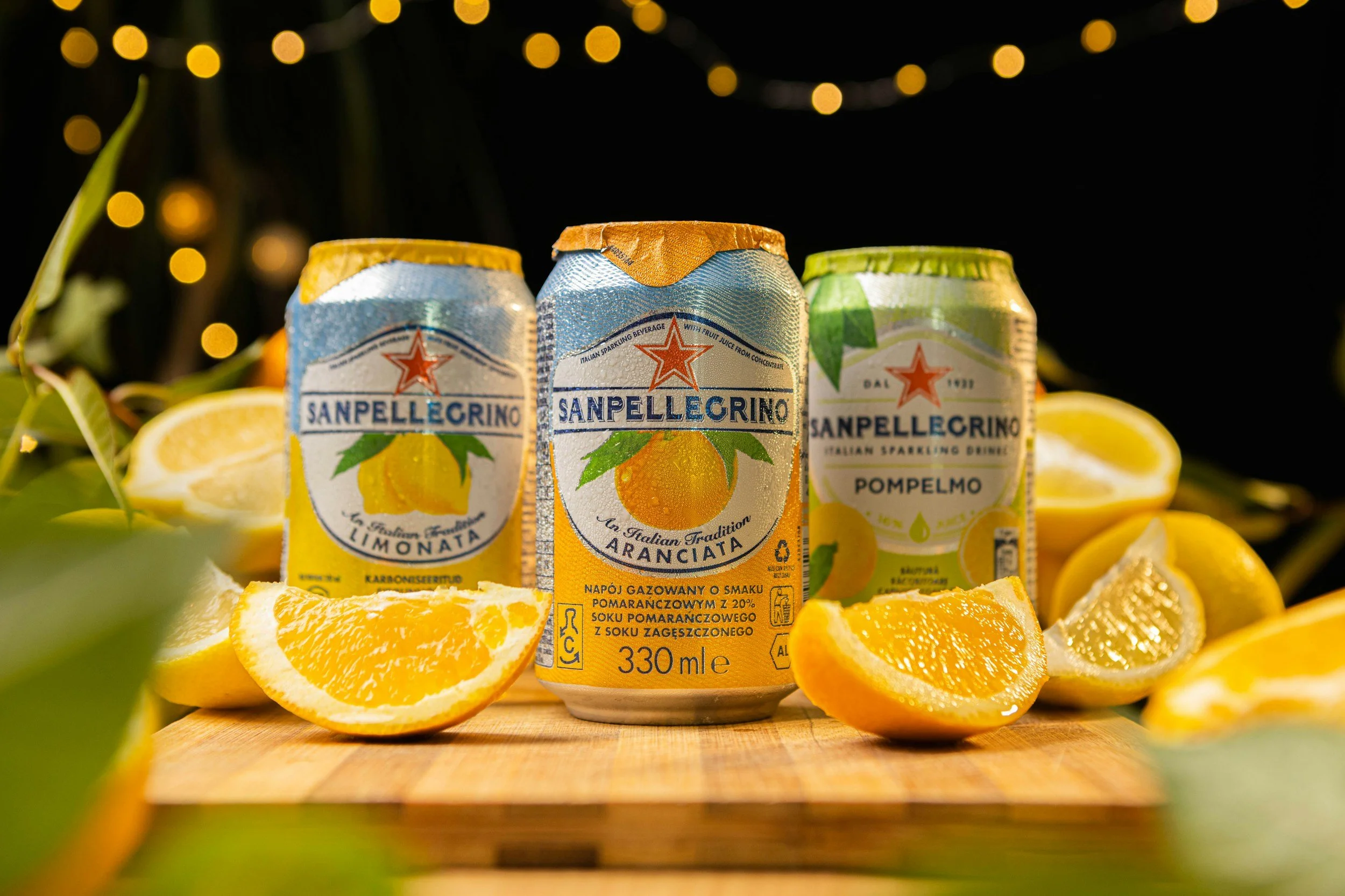 Cans of San Pellegrino sparkling drinks with lemon, orange, and pomegranate flavors surrounded by lemon slices and green leaves, with a black background and string lights above.