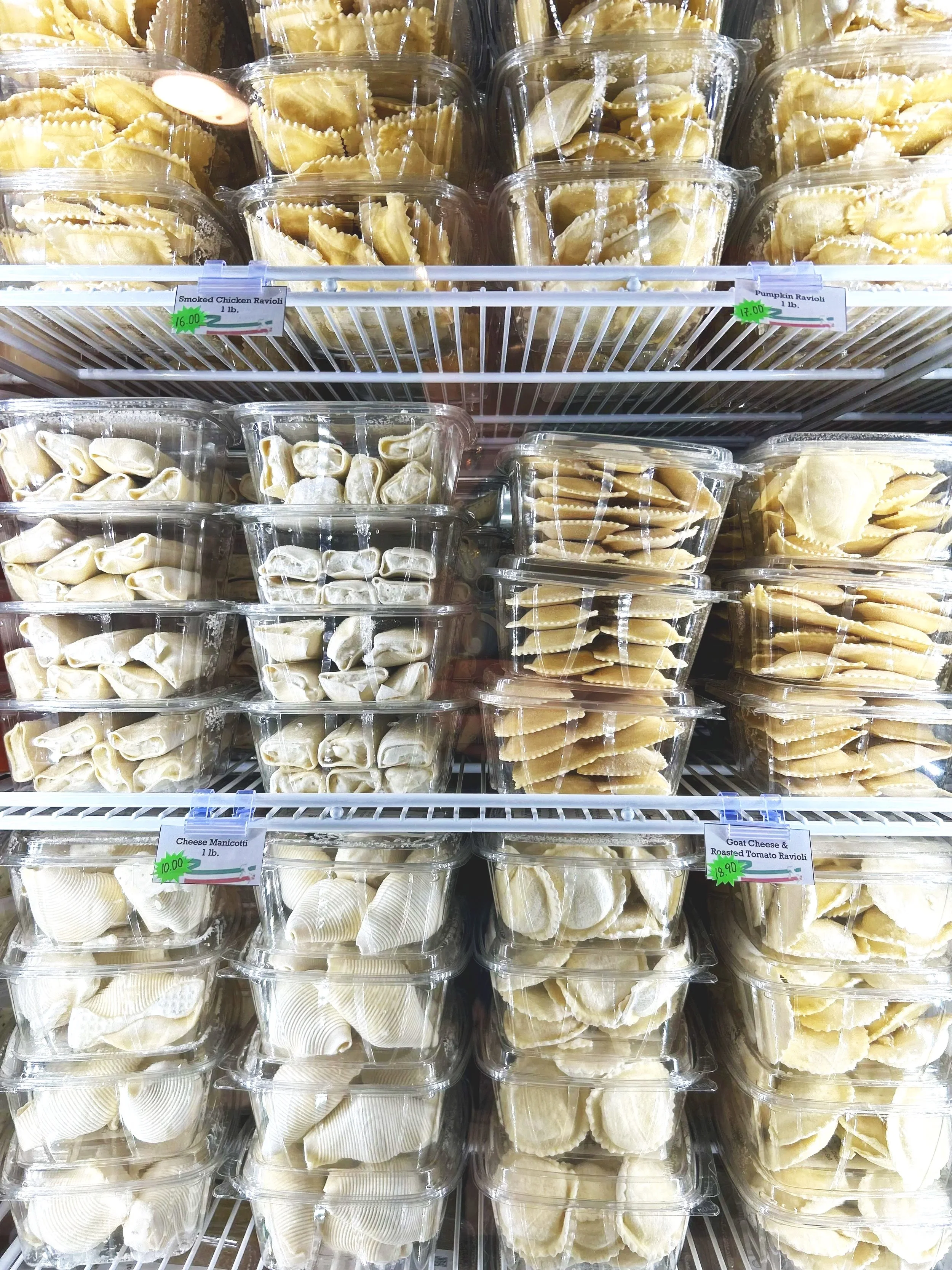 Shelves filled with clear plastic containers of ravioli pasta in a grocery store. The containers are labeled with prices and varieties such as smoked chicken, pumpkin, cheese, and goat cheese with roasted tomato. The ravioli are arranged by type in r