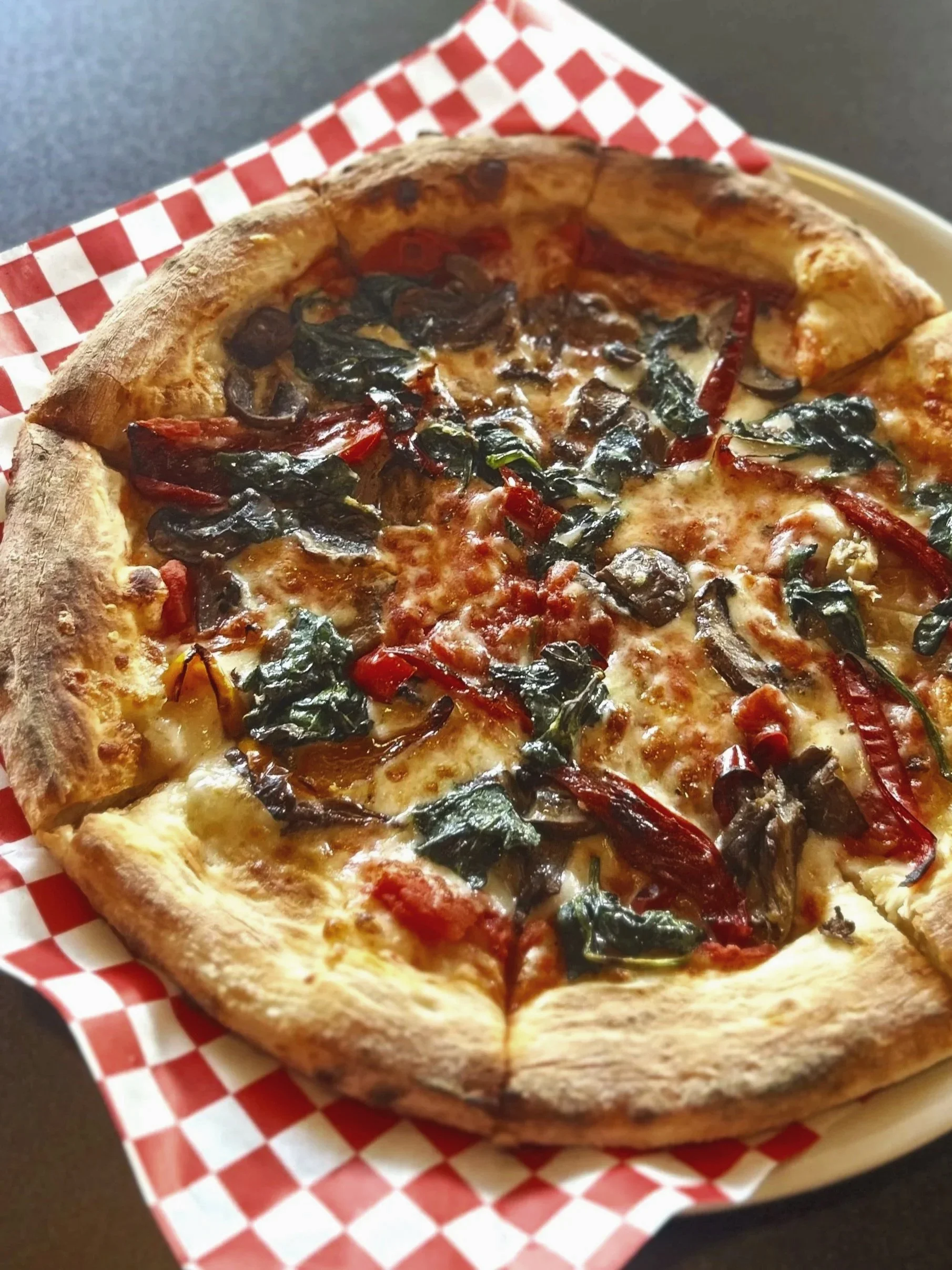 Pizza displayed on a table on top of checkered paper. Crunchy, golden crust topped with an assortment of vegetables. 