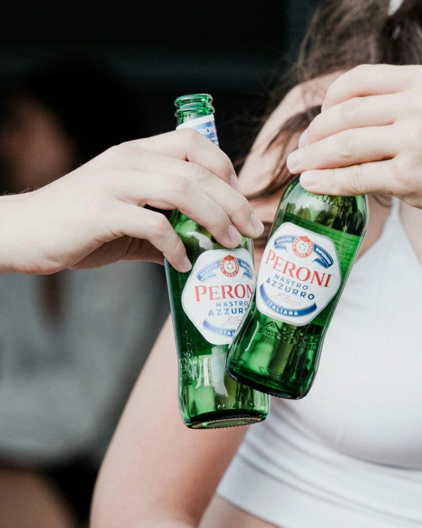 Two people holding green glass bottles of Peroni beer, clinking them together.