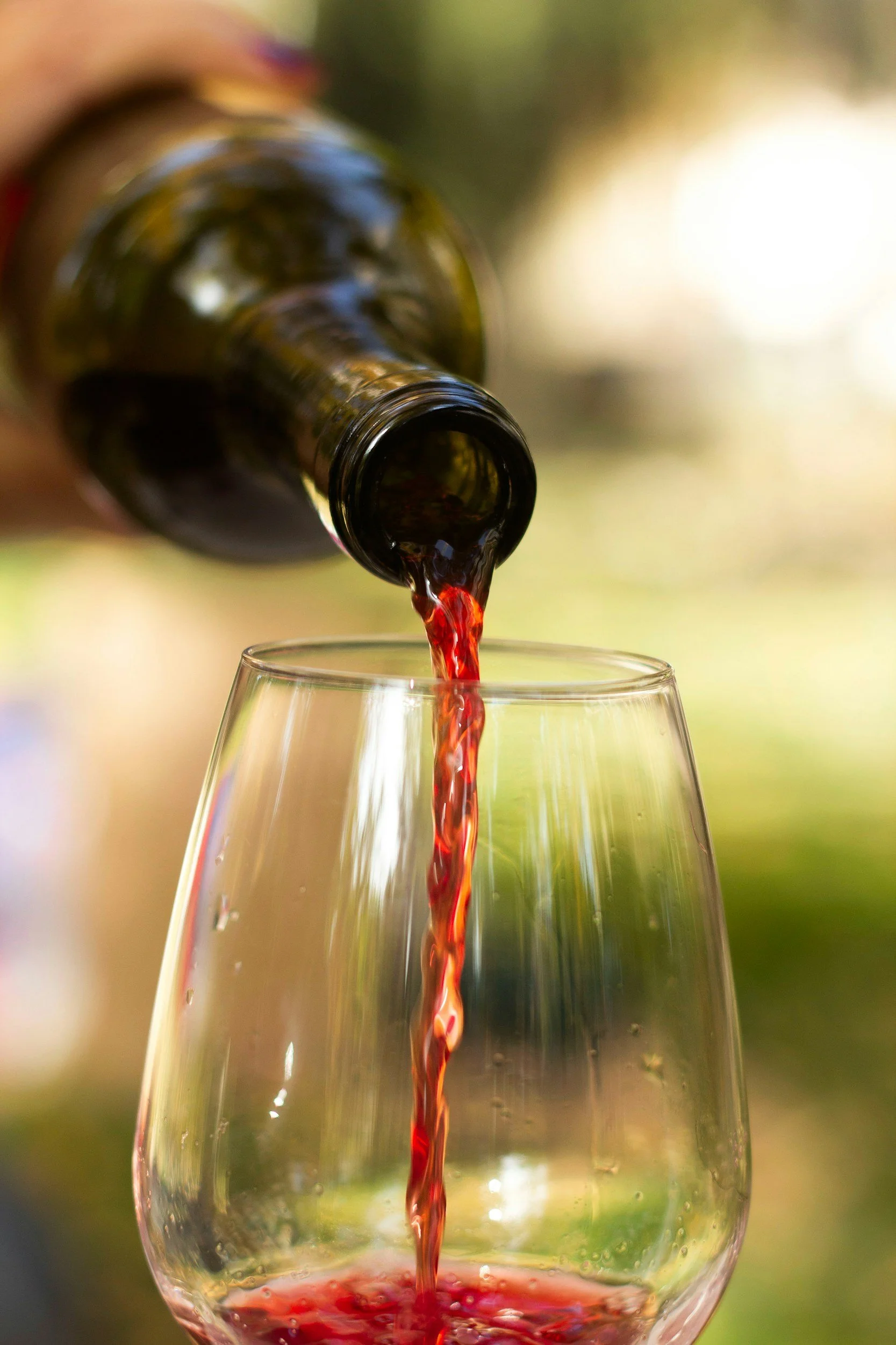 Italian red wine is being poured from a bottle into a wine glass outdoors, with a blurred natural background.