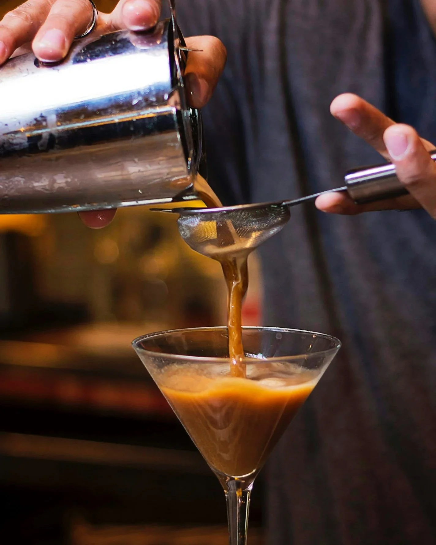 A person pouring espresso martini from a metal shaker through a strainer into a martini glass.