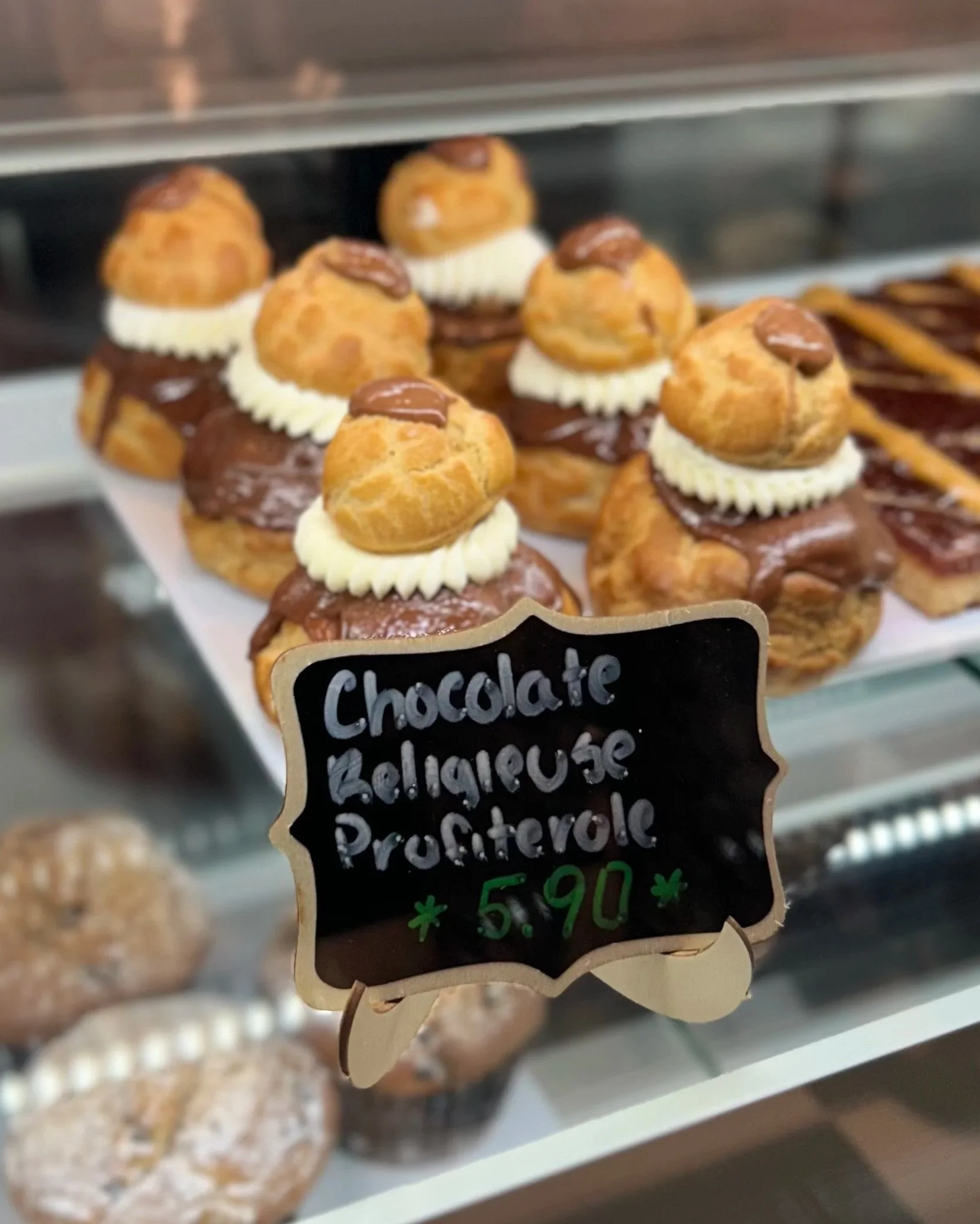 Display case with assorted pastries, including cream-filled profiteroles topped with chocolate, and a handwritten price sign indicating cost.