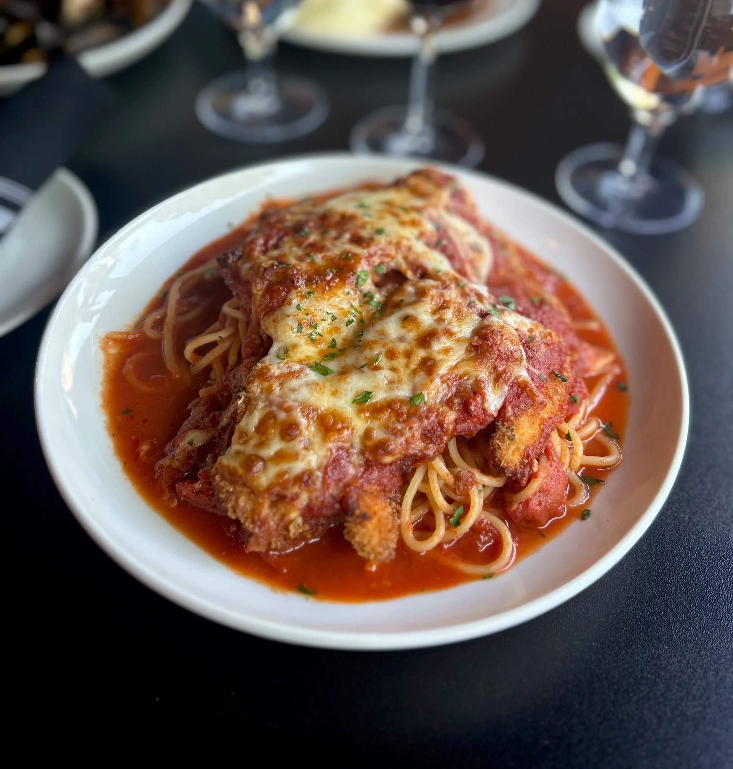 A plate of baked chicken parmesan with spaghetti, pomodoro tomato sauce, mozzarella, garnished with herbs.