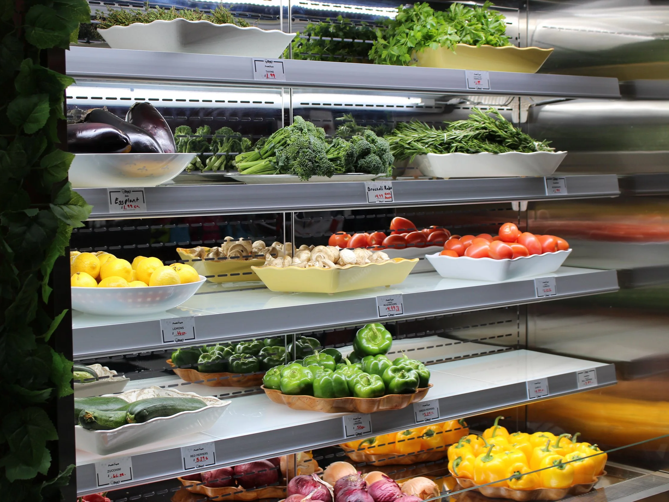 A colorful refrigerated open display of fresh produce including eggplant, lemons, garlic, onions, Roma tomatoes, fresh herbs, cucumber, broccolini, yellow and green peppers.