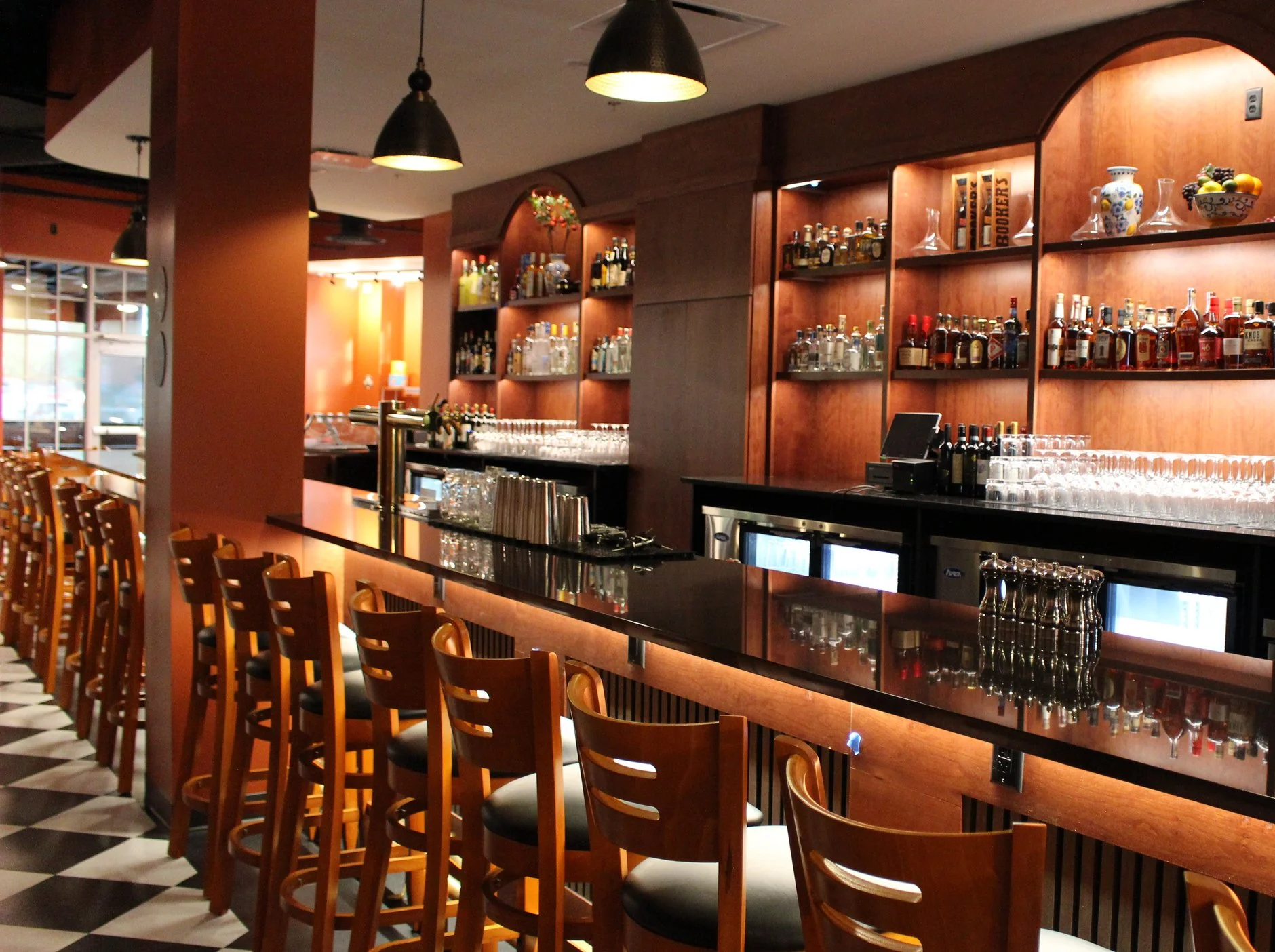 Interior of an elegant bar with wooden shelving stocked with various bottles of liquor, bowls of fruit, and glassware, illuminated by ceiling lights. There are wooden bar stools lined up along a black countertop.