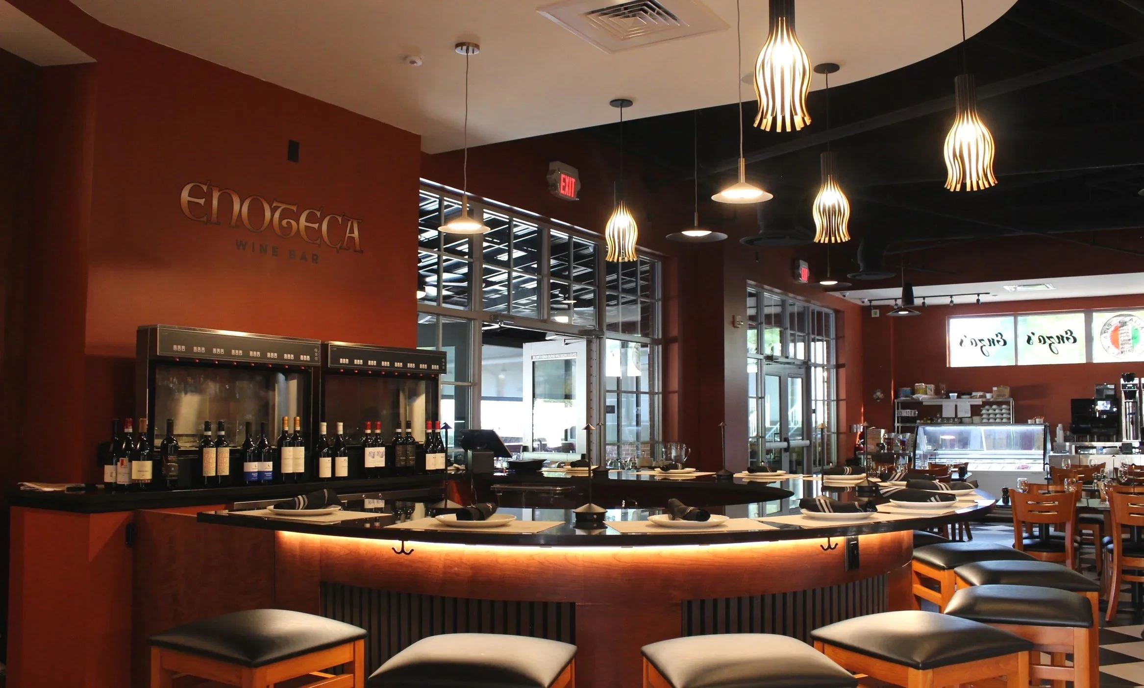 Interior of Enoteca wine bar with wine bottles on shelves and a wine fridge, hanging light fixtures, tables set with napkins and plates, and decorative greenery.