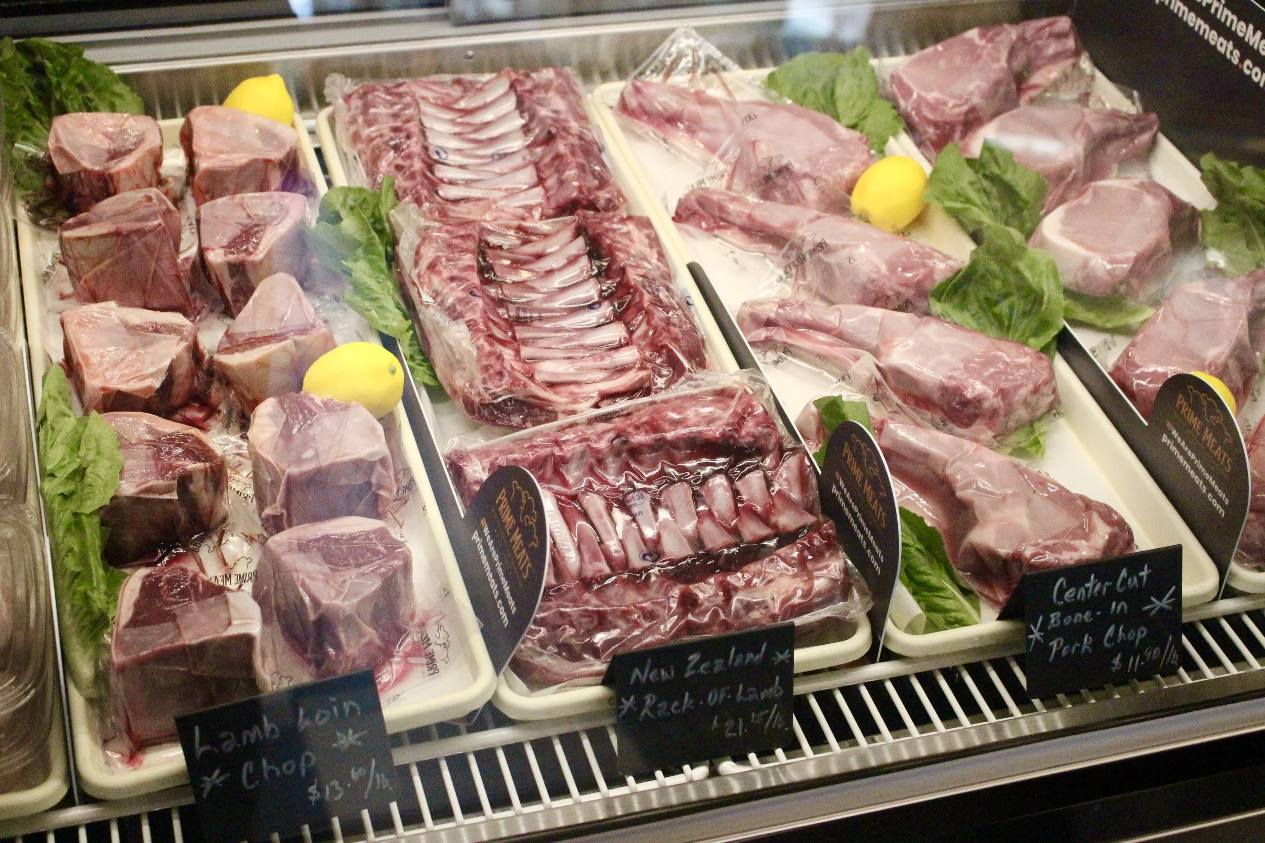 Assorted raw meats including beef and lamb cuts displayed on trays with green lettuce and yellow lemons for decoration, in a refrigerated grocery store butchery display case.