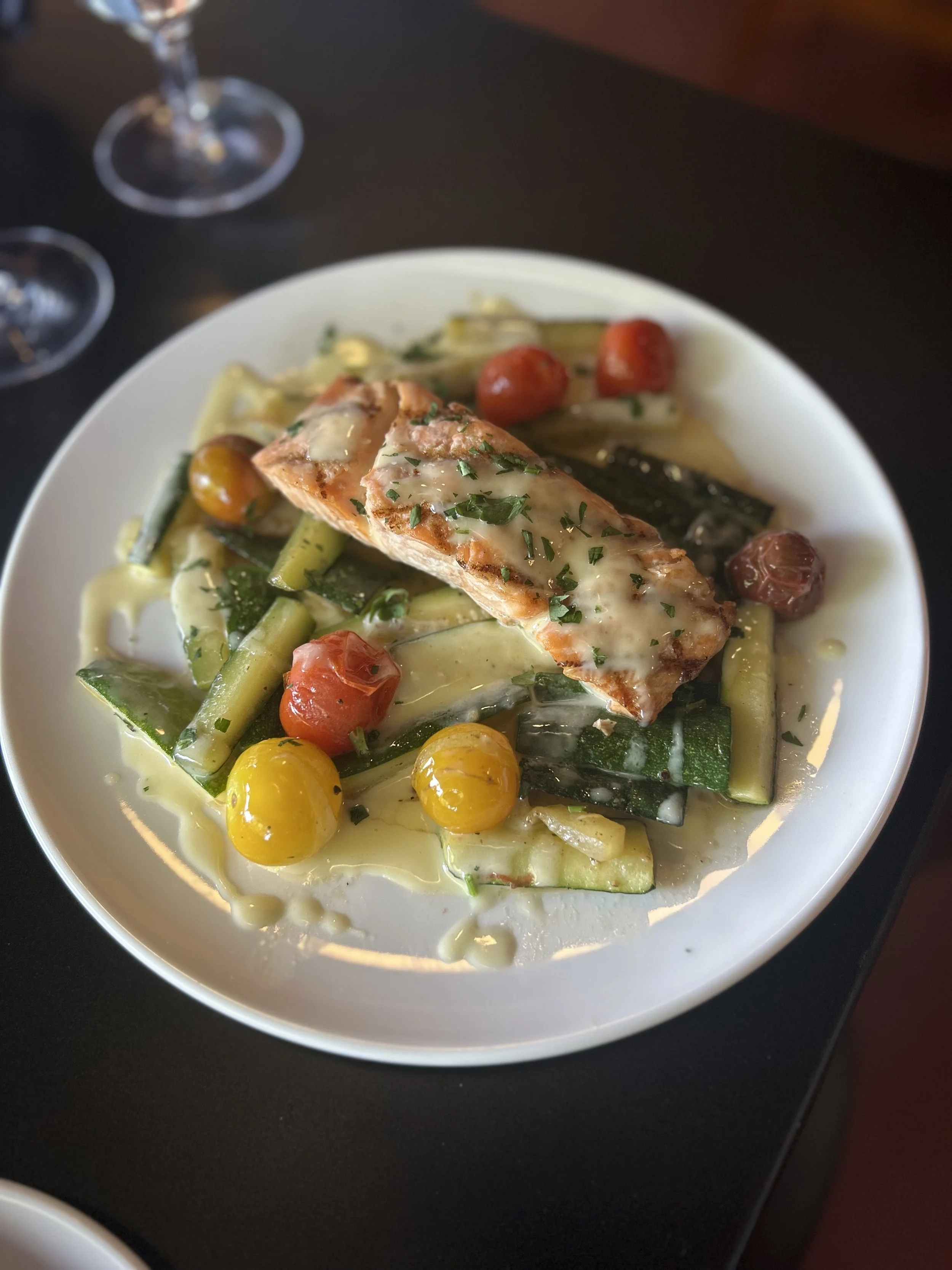 A white plate with grilled Verlasso salmon topped with beurre blanc sauce and herbs, served with zucchini, shallots, cherry tomatoes, in a light sauce.