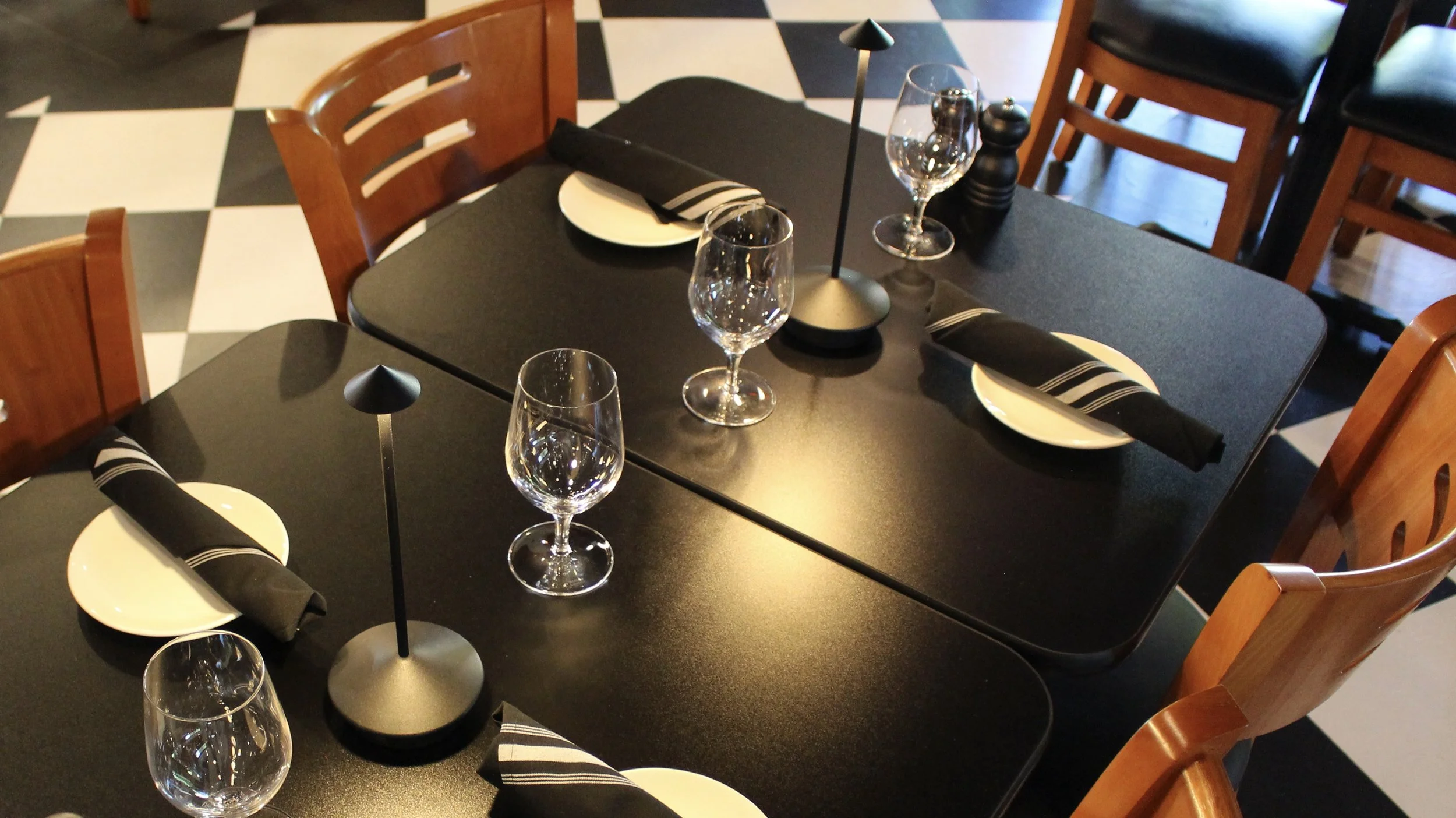 Inviting Osteria table setting with wine glasses, plates, and cloth napkins. Ambience is created by delicate table lamps on a low light setting. 