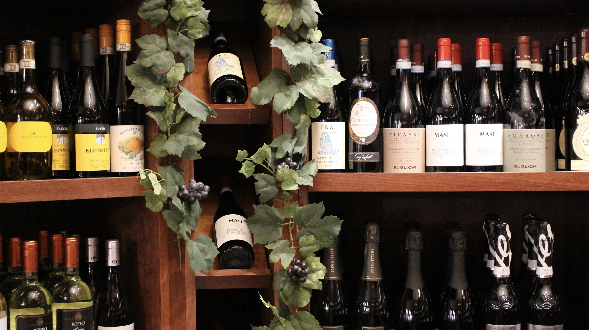 A display of wine Italian bottles on wooden shelves, decorated with green grapevine leaves and black grape clusters.