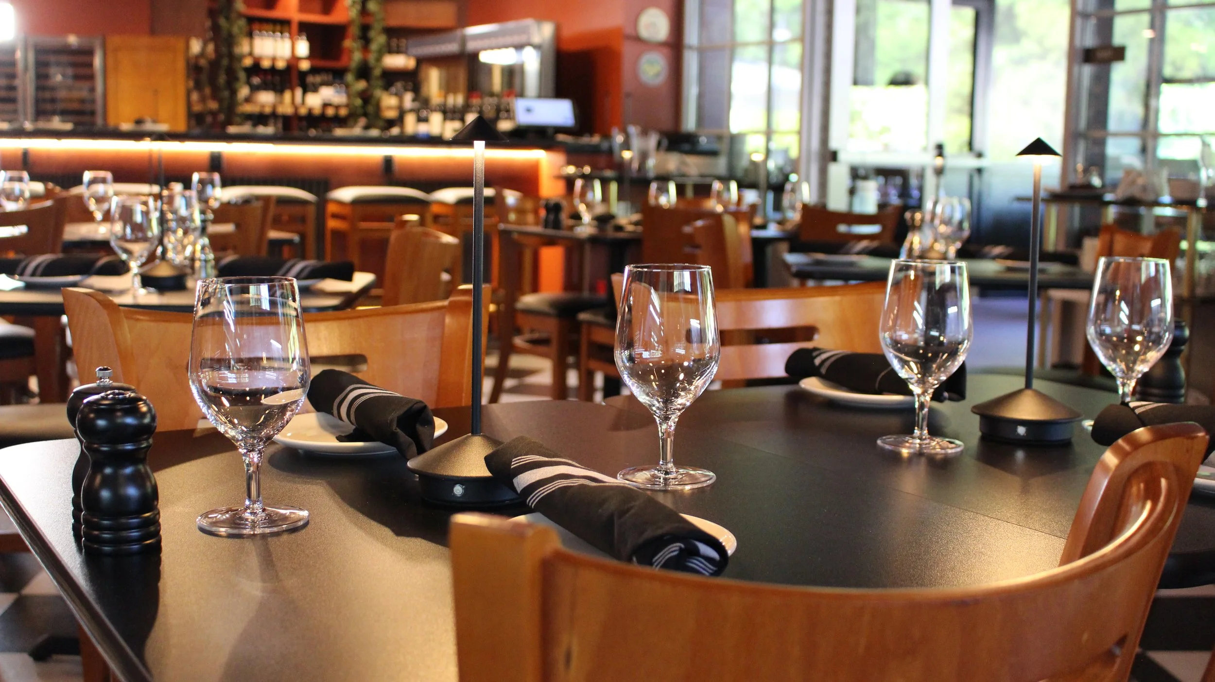 Empty restaurant dining area with tables set with wine glasses, napkins, and salt and pepper shakers, featuring large windows and wooden chairs.