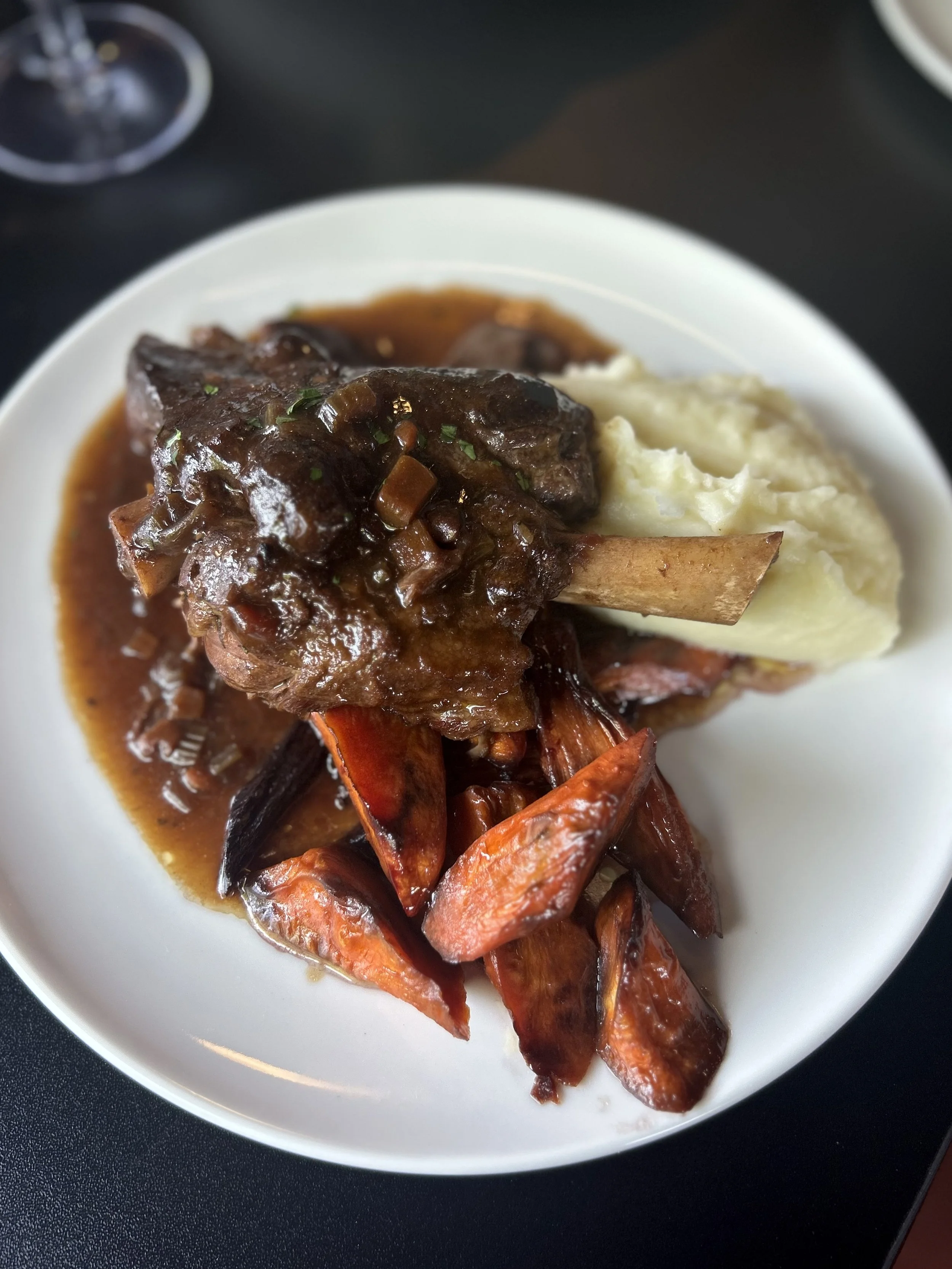 A plate of braised lamb shank with mashed potatoes, roasted carrots, topped with gravy and garnished with herbs.