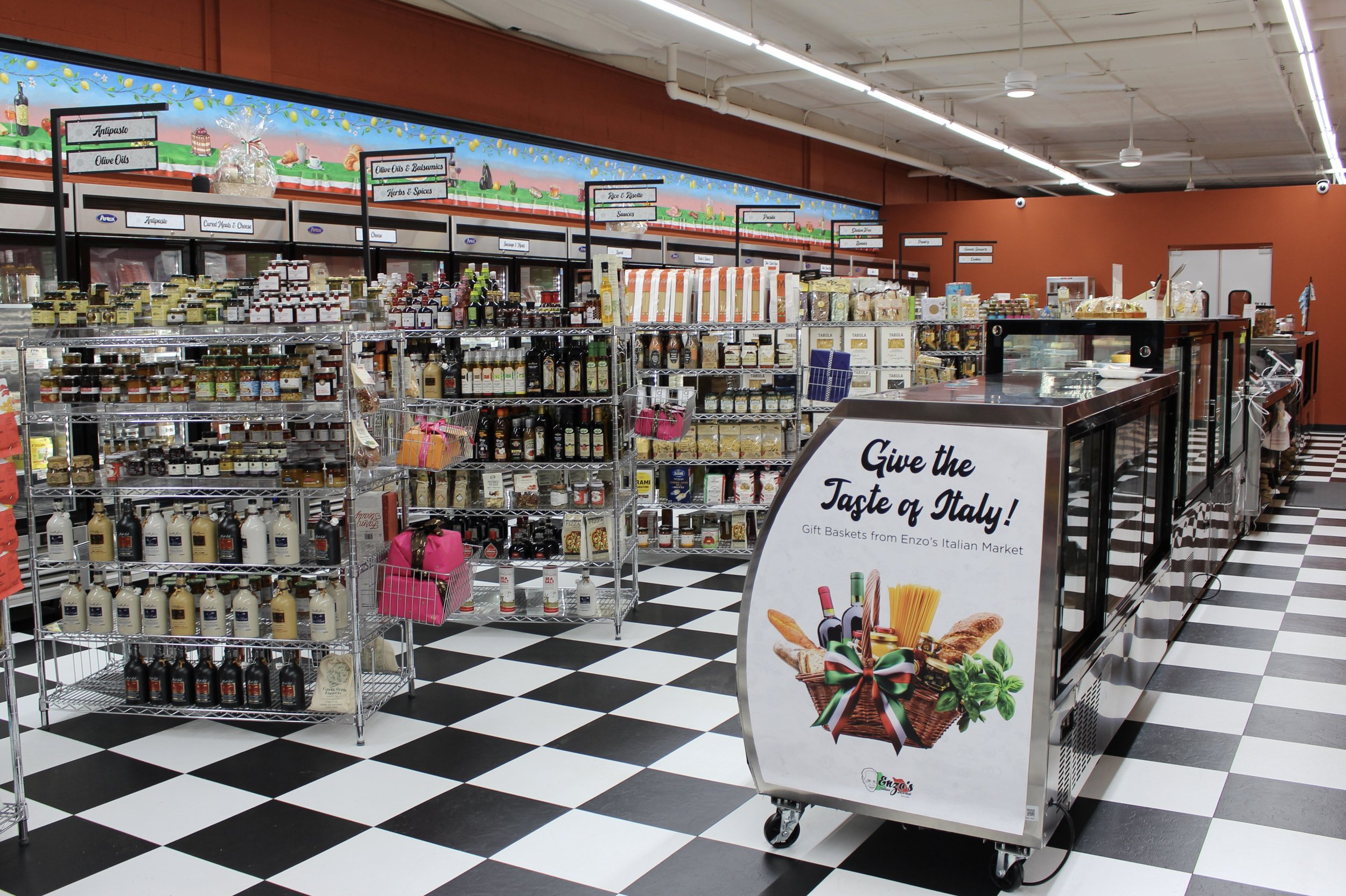 Interior of a grocery store with shelves stocked with various Italian food products, a promotional sign for Italian gift baskets, and colorful decorations.