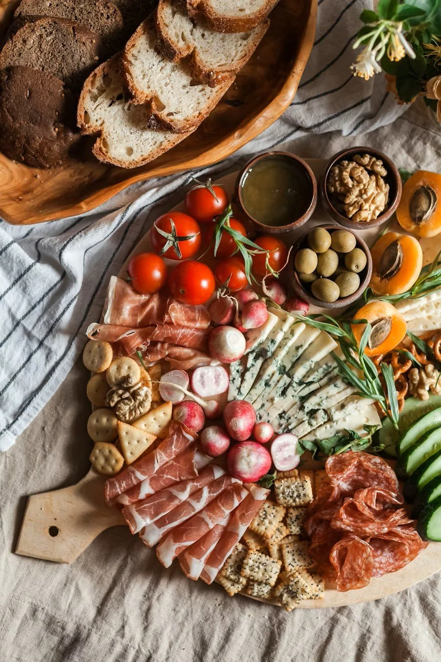 A cheese and charcuterie board with sliced bread, tomatoes, radishes, various cheeses, cured meats, nuts, olives, and grapes, arranged on a wooden serving tray and a table with a striped cloth.