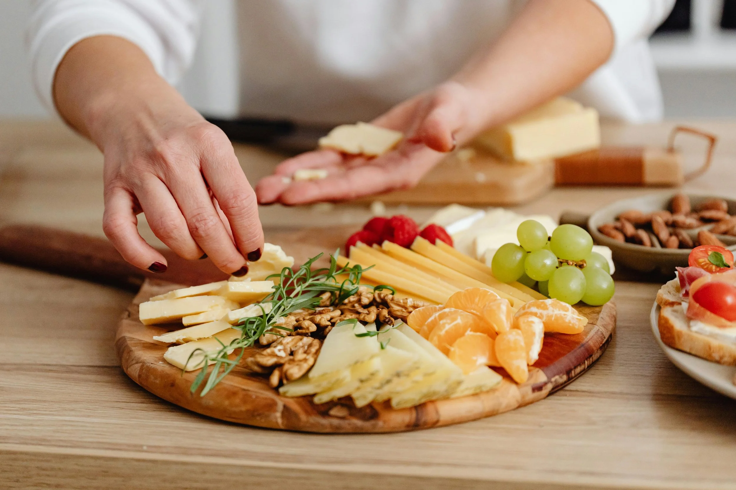 Person arranging a cheese and fruit platter with slices of cheese, grapes, raspberries, tangerine segments, walnuts, and herbs on a wooden serving board.