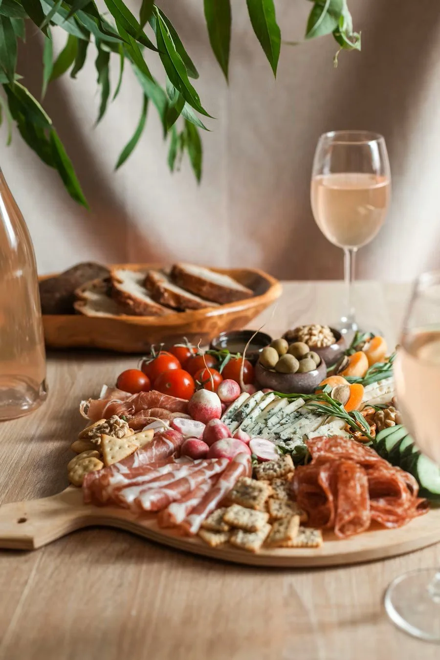 A charcuterie board with meats, cheeses, cherry tomatoes, radishes, olives, and nuts, with glasses of rosé wine and a bowl of bread slices in the background, set on a wooden table with a green leafy plant overhead.
