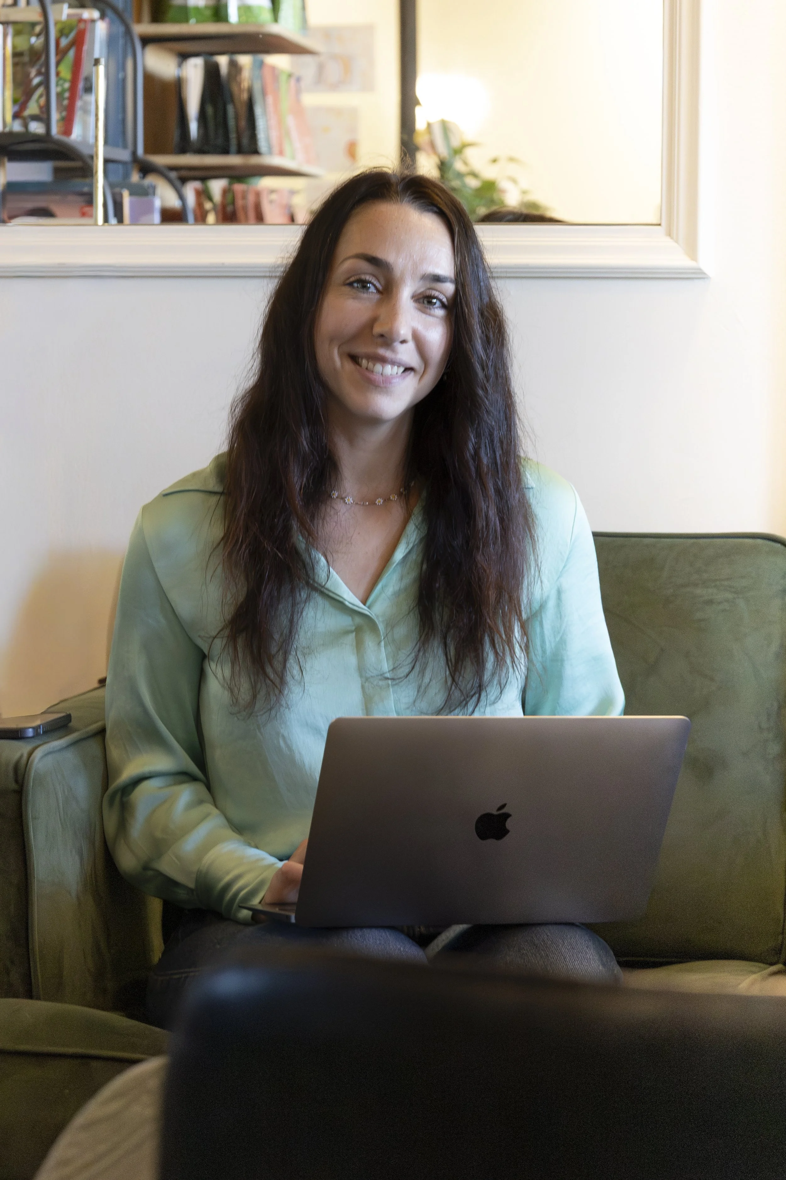 Une femme souriante avec des cheveux longs bruns, portant un haut vert pastel, assise sur un canapé vert avec un ordinateur portable Apple sur ses genoux, dans un intérieur avec une bibliothèque en arrière-plan.