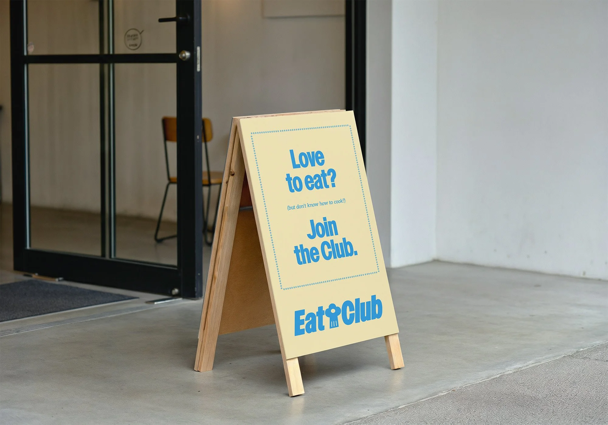 Photograph of an a-frame sign outside of a modern open-plan space. The sign reads "Love to eat? (but don't know how to cook?) Join the Club." Below the text is the EatClub logo.