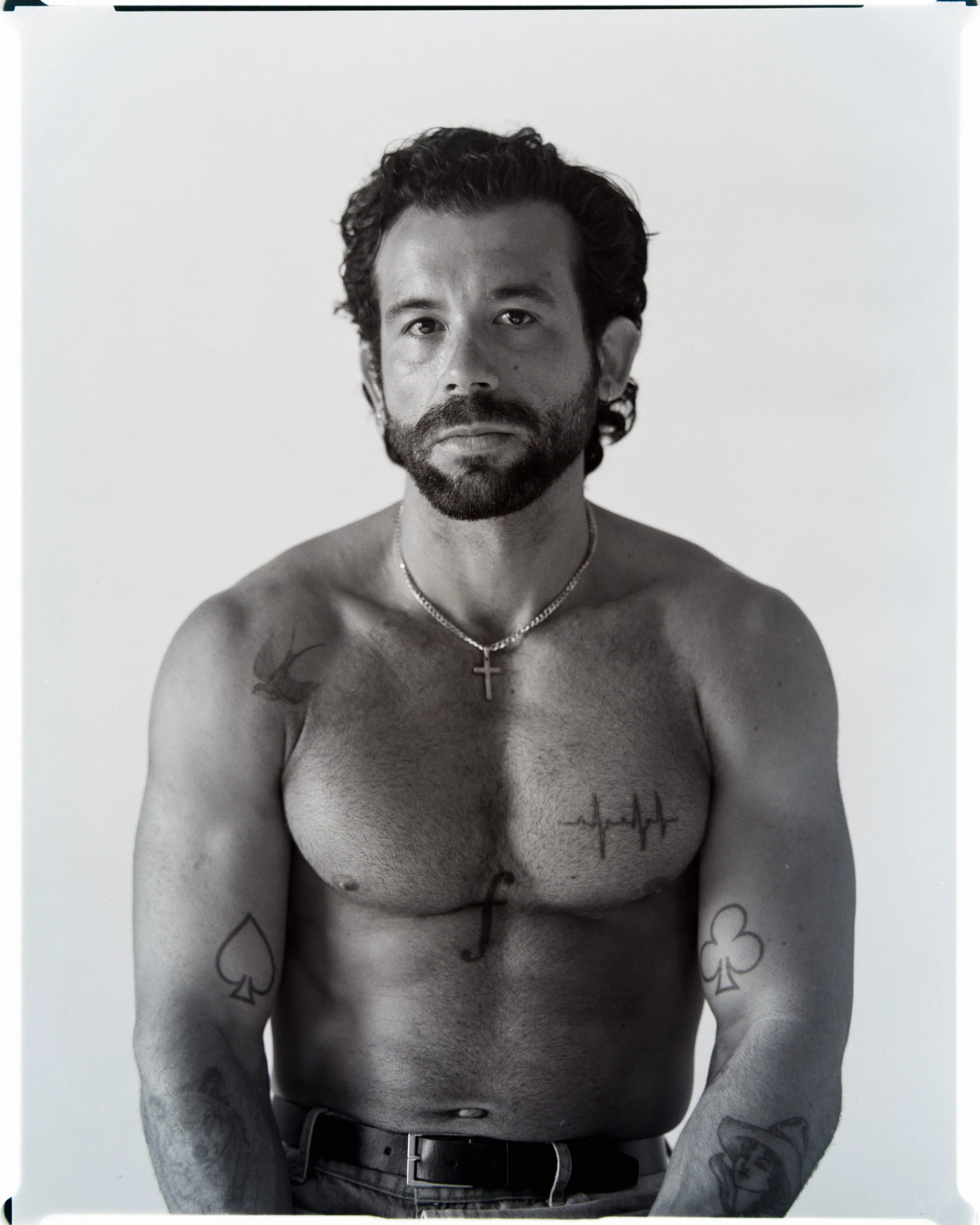 A shirtless man with tattoos on his arms and chest, wearing a necklace with a cross, poses against a plain background.