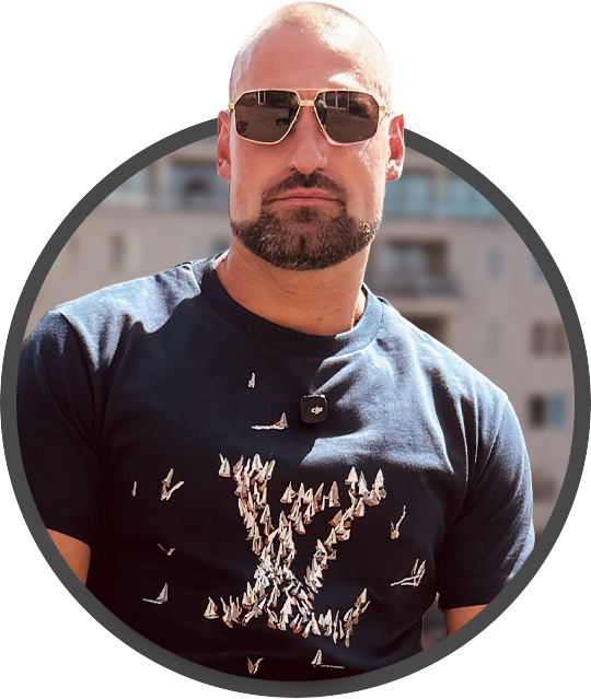 Oscar Strong sunglasses and beard wearing a dark blue t-shirt with a butterfly design, standing outdoors against a blurred cityscape background.