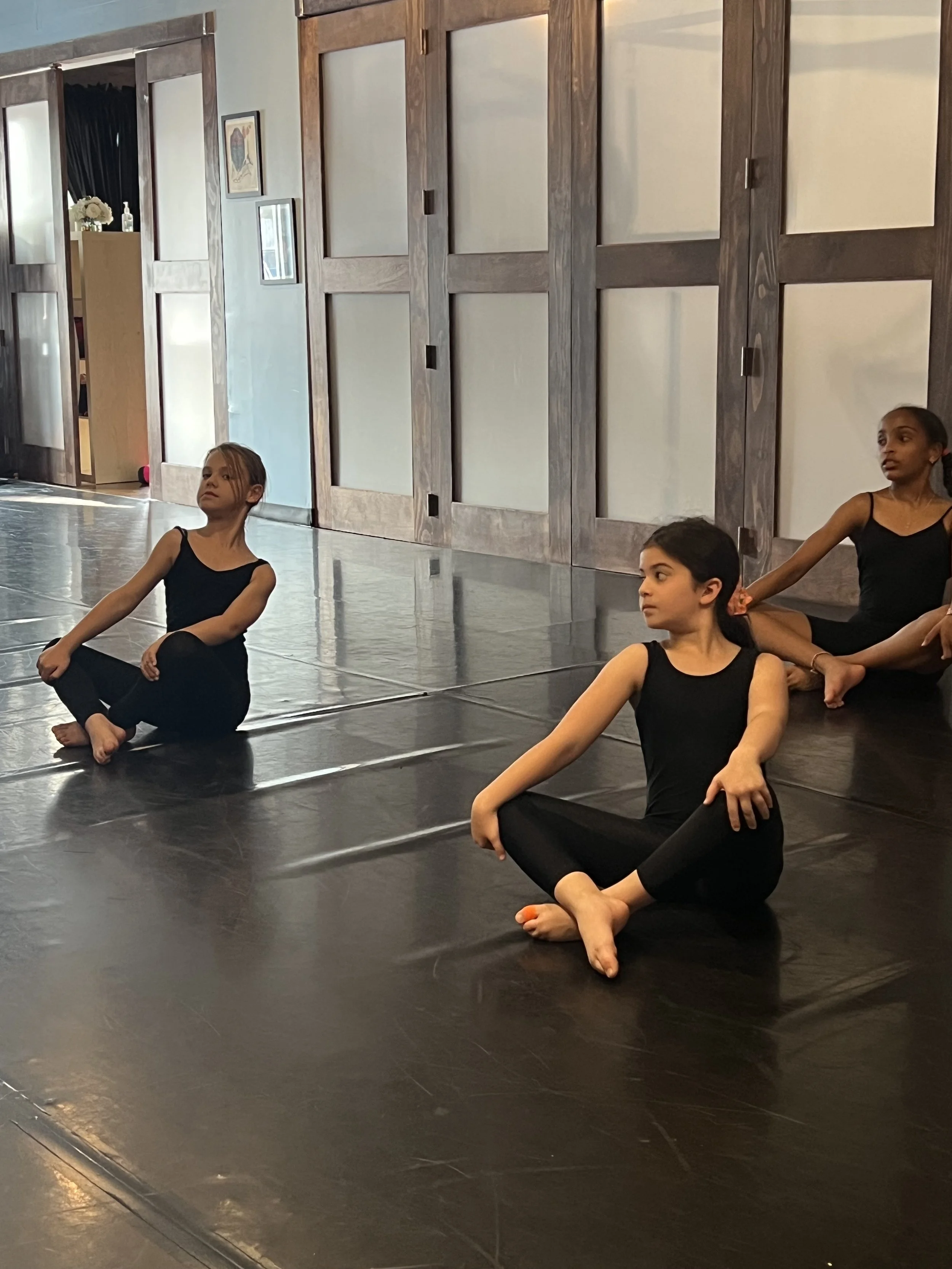Three young girls in black leotards sitting on a polished studio floor during a ballet class or practice.